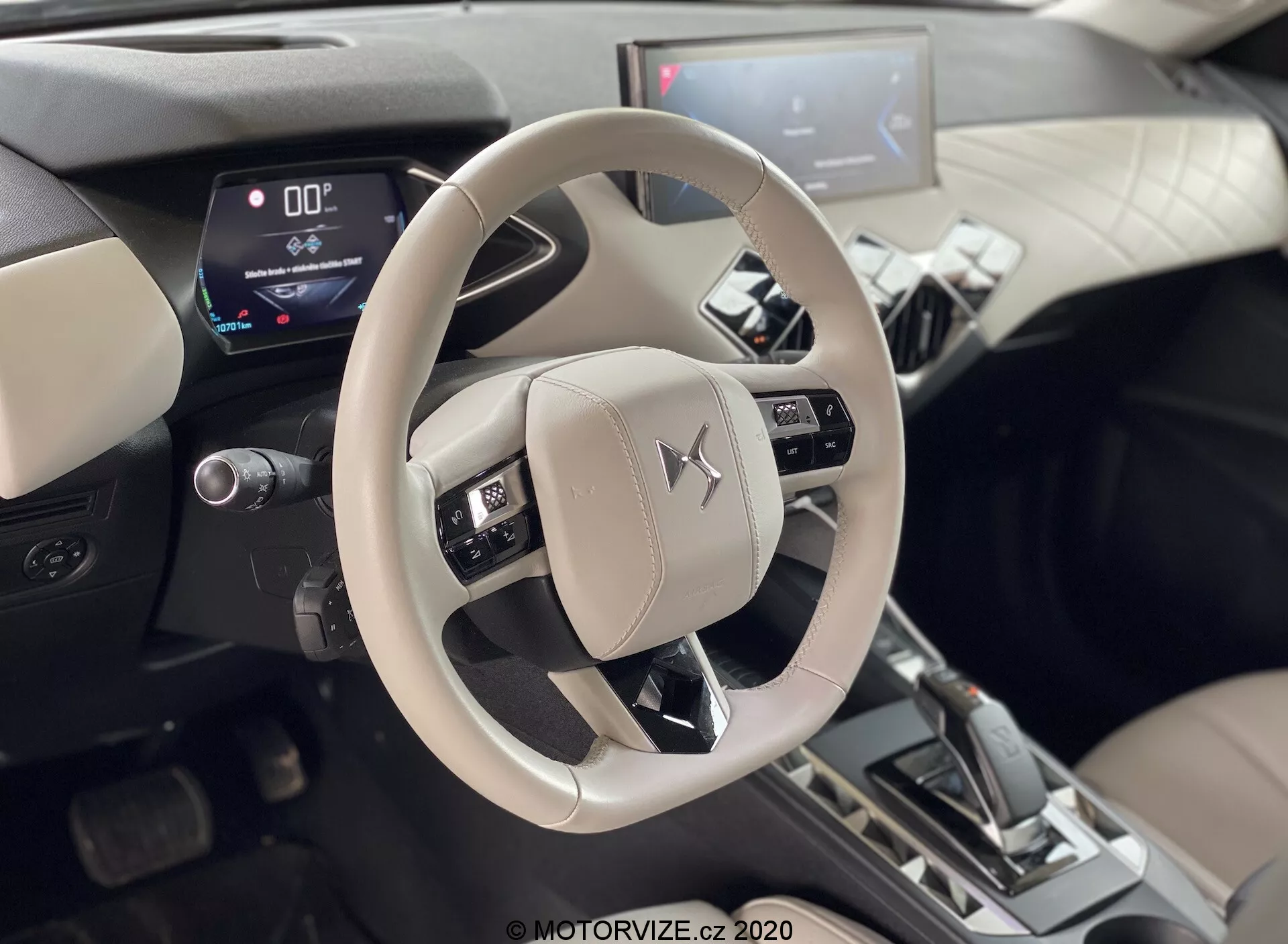 Vue intérieure du DS-Automobiles DS-3 2019 (DS-3 Crossback E-Tense) centrée sur le volant beige clair portant l'emblème Mitsubishi et la console centrale. Pris de la perspective des sièges arrière, présentant les commandes montées pour l'audio, la voix et les systèmes de contrôle de croisière. Tableau de bord numérique affichant une vitesse de zéro et levier de vitesse en position 'P', indiquant que la voiture est stationnée. Système d'infodivertissement central avec écran tactile visible dans un intérieur bicolore avec accents métalliques, suggérant des affichages numériques modernes et des fonctionnalités automobiles avancées.