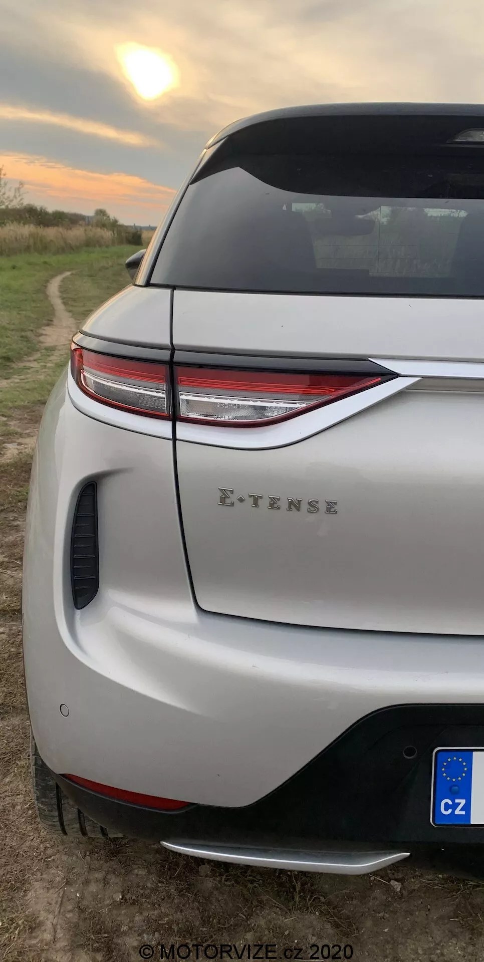 Gros plan vertical de la section arrière du DS-3 2019 (DS-3 Crossback E-TENSE). Accent sur les feux arrière LED modernes, le pare-chocs arrière sportif avec diffuseur intégré et capteur de stationnement, le couvercle de coffre avec spoiler aérodynamique et vue partielle de la roue arrière et de l'aile. Cadre rural avec un champ ouvert sous un ciel de lever ou de coucher de soleil, indiquant la conception moderne du véhicule et ses capacités électriques.