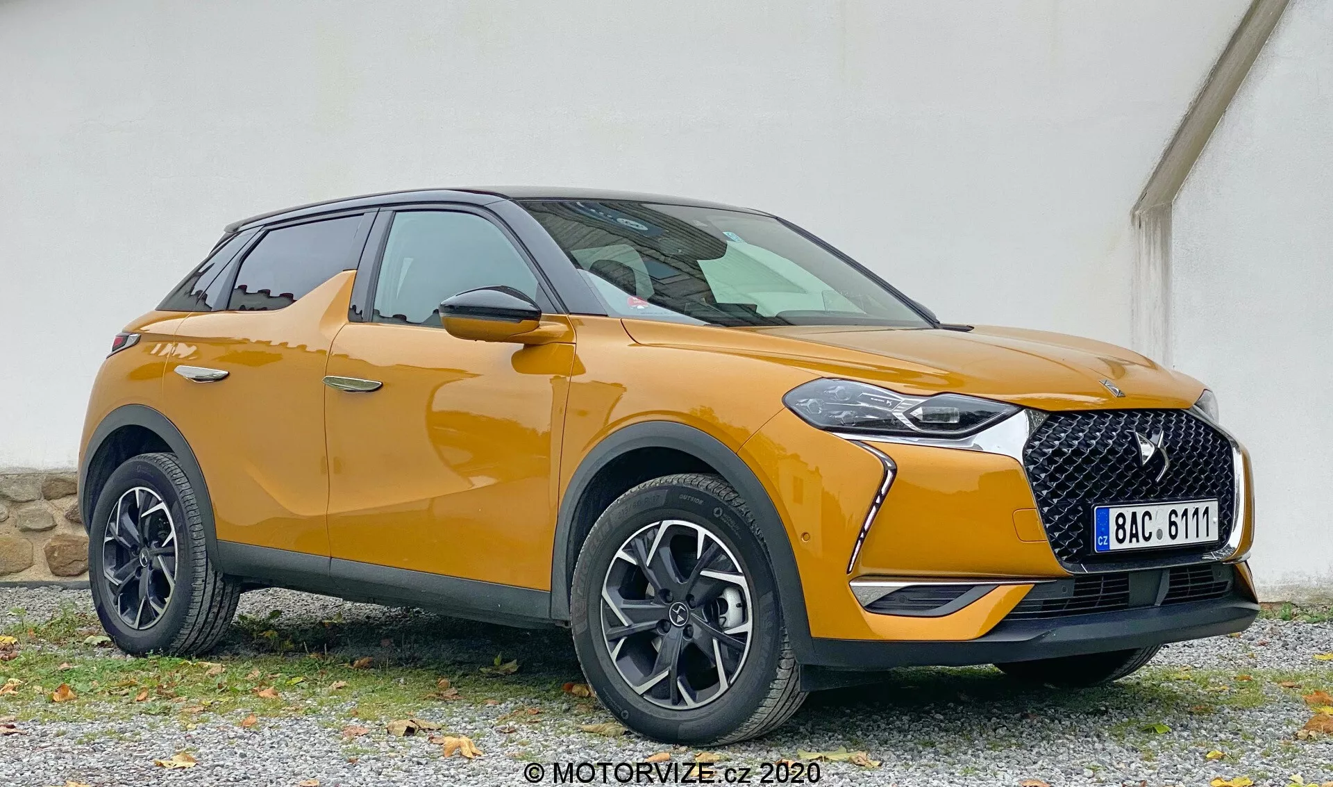 SUV moderne en vue de trois quarts avant montrant la DS Automobiles DS 3 2019 (DS 3 Crossback) avec des détails du coin avant gauche, y compris le phare, la calandre, le capot, le pare-chocs avant, la roue, l'aile avant et la porte du conducteur, positionné sur une surface en gravier contre un fond de mur de couleur atténuée.
