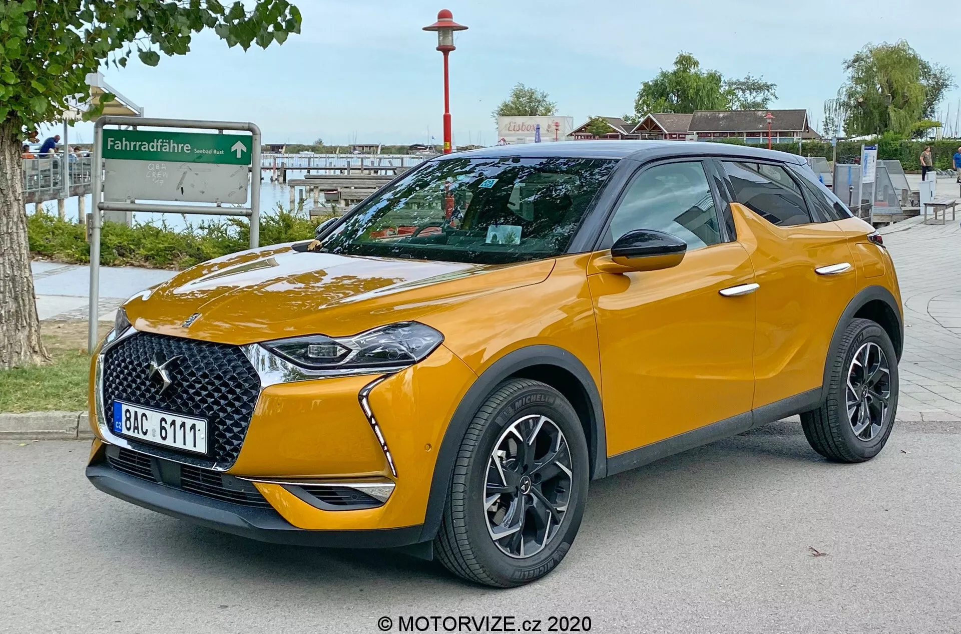 Image montrant le SUV moderne DS Automobiles DS 3 Crossback 2019 en vue de trois quarts avant, mettant en avant la grille avant élégante, les phares LED intégrés, le pare-chocs avant, le capot et le profil du côté conducteur. Garé à l'extérieur sur une surface pavée avec un arrière-plan urbain comprenant une station de partage de vélos. La photo, prise d'un point de vue bas au niveau des phares, accentue la garde au sol de la voiture. Les caractéristiques visibles incluent des roues en alliage à design multi-branches, des rétroviseurs avec clignotants intégrés, des pneus à profil bas, une couleur de toit contrastante avec des barres de toit, une finition de peinture métallisée, des badges spécifiques au trim et des vitres arrière teintées. La plaque d'immatriculation est intentionnellement floutée pour la confidentialité.