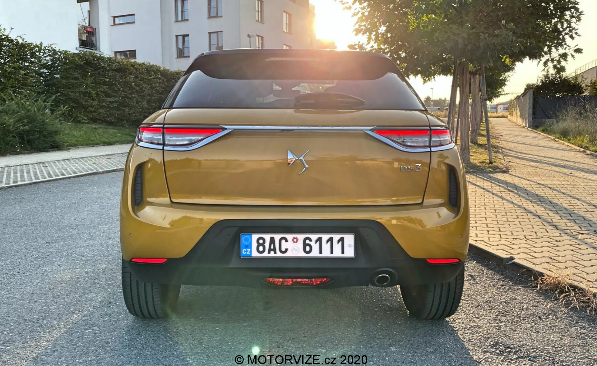 Vue arrière d'une voiture DS Automobiles DS 3 2019 (DS 3 Crossback) de couleur dorée à l'aube ou au crépuscule. L'image montre les éléments de design arrière de la voiture, y compris un emblème en forme d'étoile et les chiffres '300' sur le couvercle du coffre, des feux arrière à LED horizontaux, une plaque d'immatriculation de style européen, un pare-chocs arrière avec des réflecteurs et des capteurs de stationnement, un diffuseur et une finition en peinture métallique.