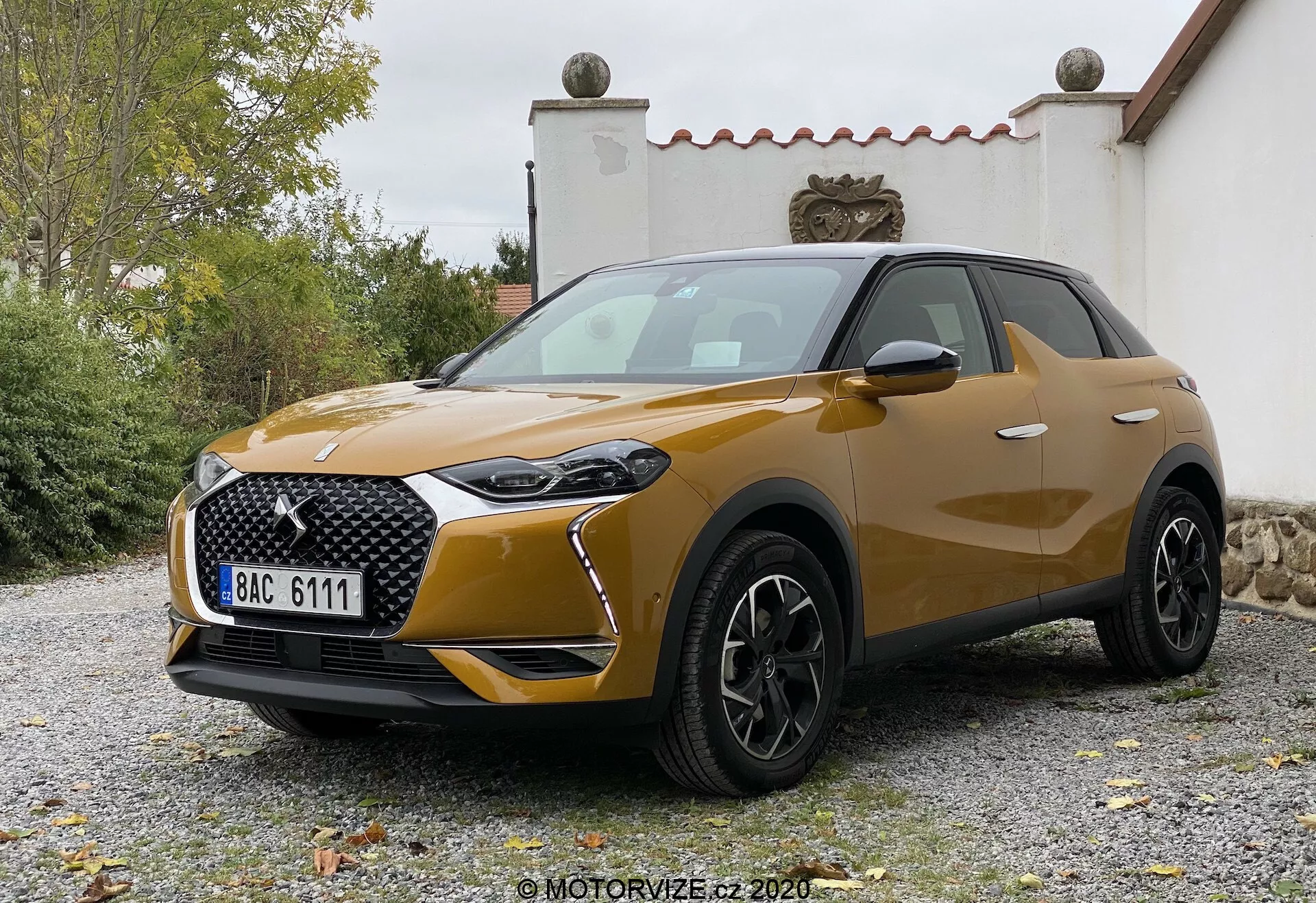 Vue de trois quarts avant de la DS-3 Crossback 2019, un crossover SUV compact moderne. L'angle légèrement abaissé met en valeur sa hauteur et son allure robuste, sous un éclairage doux et diffus. Les caractéristiques clés incluent des feux de jour à LED distinctifs autour du groupe principal de phares, une grande grille à motifs avec un badge proéminent, des sections de phares antibrouillard noircies dans le pare-chocs et une prise d'air notable sous la grille. Le capot présente des contours subtils, les roues sont des alliages multibranches noirs suggérant un niveau de finition supérieur, et les rétroviseurs extérieurs de couleur carrosserie, probablement avec des clignotants intégrés. Les lignes de carrosserie sont fluides et les légers renflements au-dessus des arches des roues ajoutent à la robustesse et à la sportivité. La peinture métallique indique un niveau premium. Le véhicule pourrait posséder une technologie d'éclairage avancée, des capteurs de stationnement et peut-être des systèmes de caméra ou de radar intégrés, bien que non clairement visibles. Positionné sur une surface gravillonnée avec de la verdure et une structure de bâtiment en arrière-plan, la plaque d'immatriculation de style européen est visible mais modifiée.