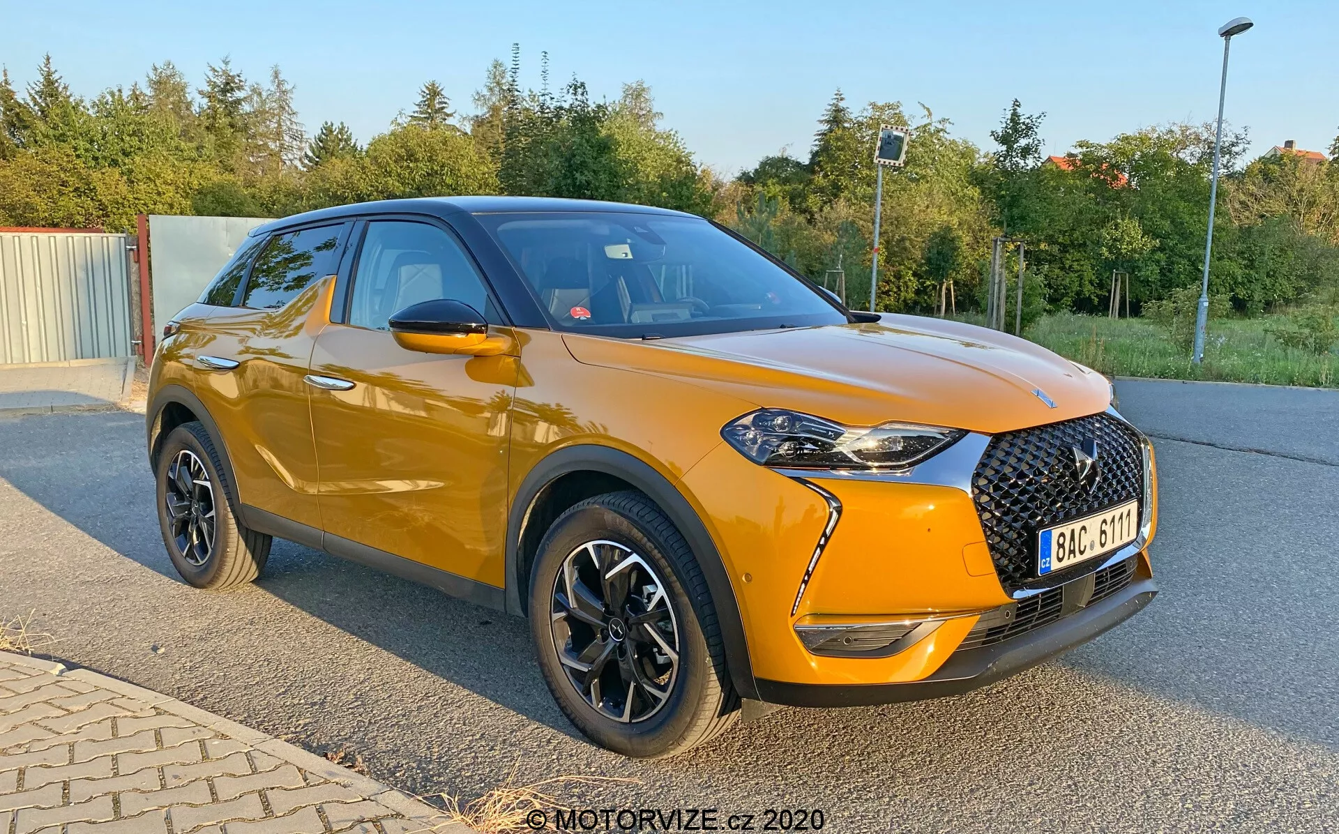 SUV compact et moderne DS Automobiles DS-3 Crossback de 2019, montré en stationnement en angle avec un accent sur son profil avant et latéral. L'image, capturée d'une perspective basse près des moyeux de roue, met en évidence la carrosserie métallisée orange vif du véhicule et le toit noir. L'avant présente une grande calandre noire à motif diamant, des phares aigus avec des accents LED et des antibrouillards dans des boîtiers noirs. La voiture dispose de rétroviseurs latéraux noirs, de garnitures de fenêtre chromées et de roues noires avec des accents argentés. Remarquez la posture légèrement surélevée, les lignes sportives de la carrosserie et le pli vers l'avant à travers les poignées des portes.