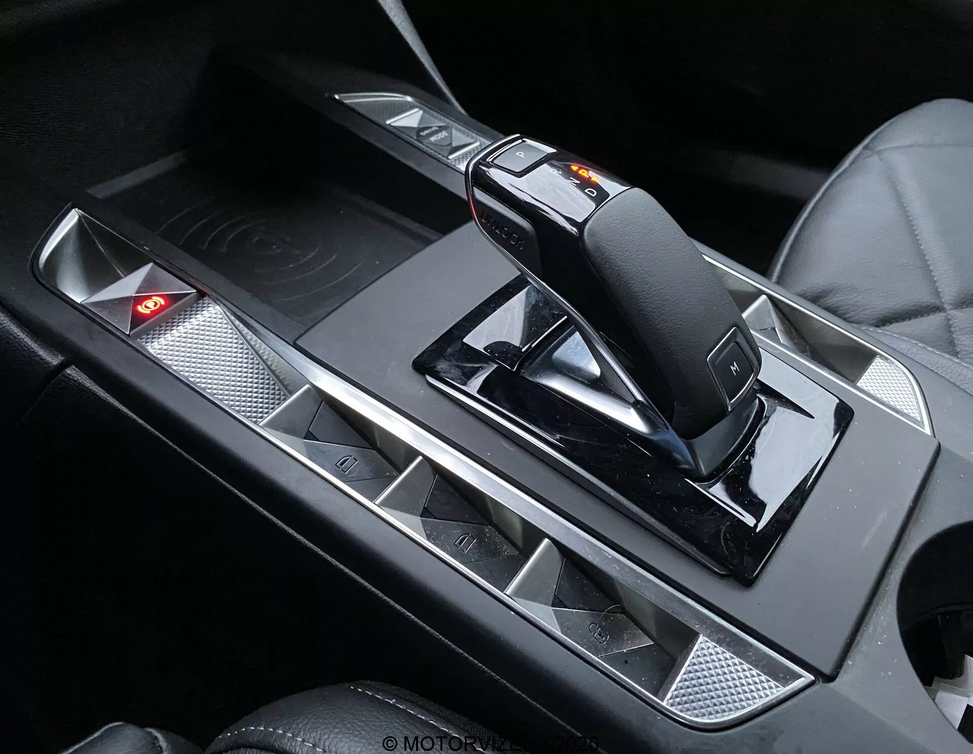 Gros plan de l'intérieur du DS Automobiles DS-3 Crossback 2019, mettant en évidence la console centrale avec son levier de vitesses moderne et ergonomique, son commutateur de mode manuel et son mécanisme de déverrouillage. La console présente des accents métalliques contrastant avec des surfaces noires, des porte-gobelets avec finition mate et un panneau de commande avec un voyant rouge allumé.