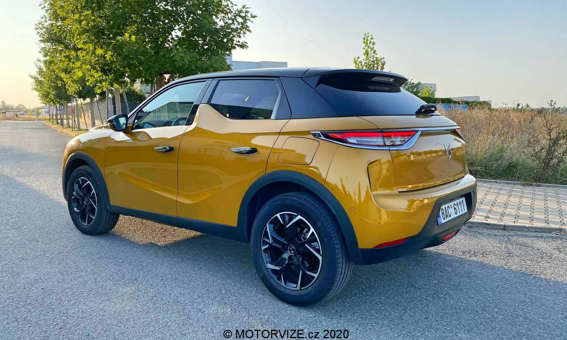 Vue arrière trois quarts d'un SUV crossover moderne en plein jour, avec une couleur or métallique vibrante et un toit noir sportif, feux arrière à LED modernes, grandes jantes en alliage noires, hauteur de caisse élevée, capteurs de stationnement arrière visibles et design bicolore, indicatif d'un DS Automobiles DS 3 Crossback 2019.