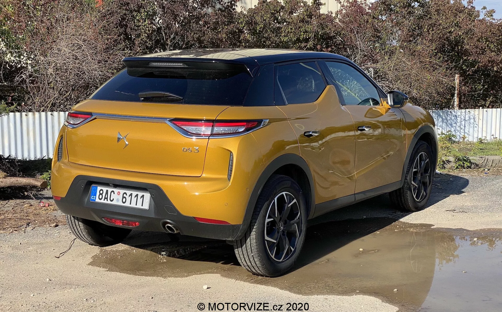 Une vue trois-quarts arrière d'un SUV DS-3 Crossback de couleur or métallique stationné en extérieur par une journée ensoleillée. L'image capture le véhicule sous un angle légèrement droit, mettant en valeur le côté arrière droit et la zone du coffre. Les caractéristiques principales incluent des feux arrière angulaires modernes, une lunette arrière inclinée avec un spoiler intégré, un pare-chocs proéminent avec des capteurs de stationnement, des roues en alliage multi-branchées noires, une garde au sol élevée et des vitres arrière teintées.