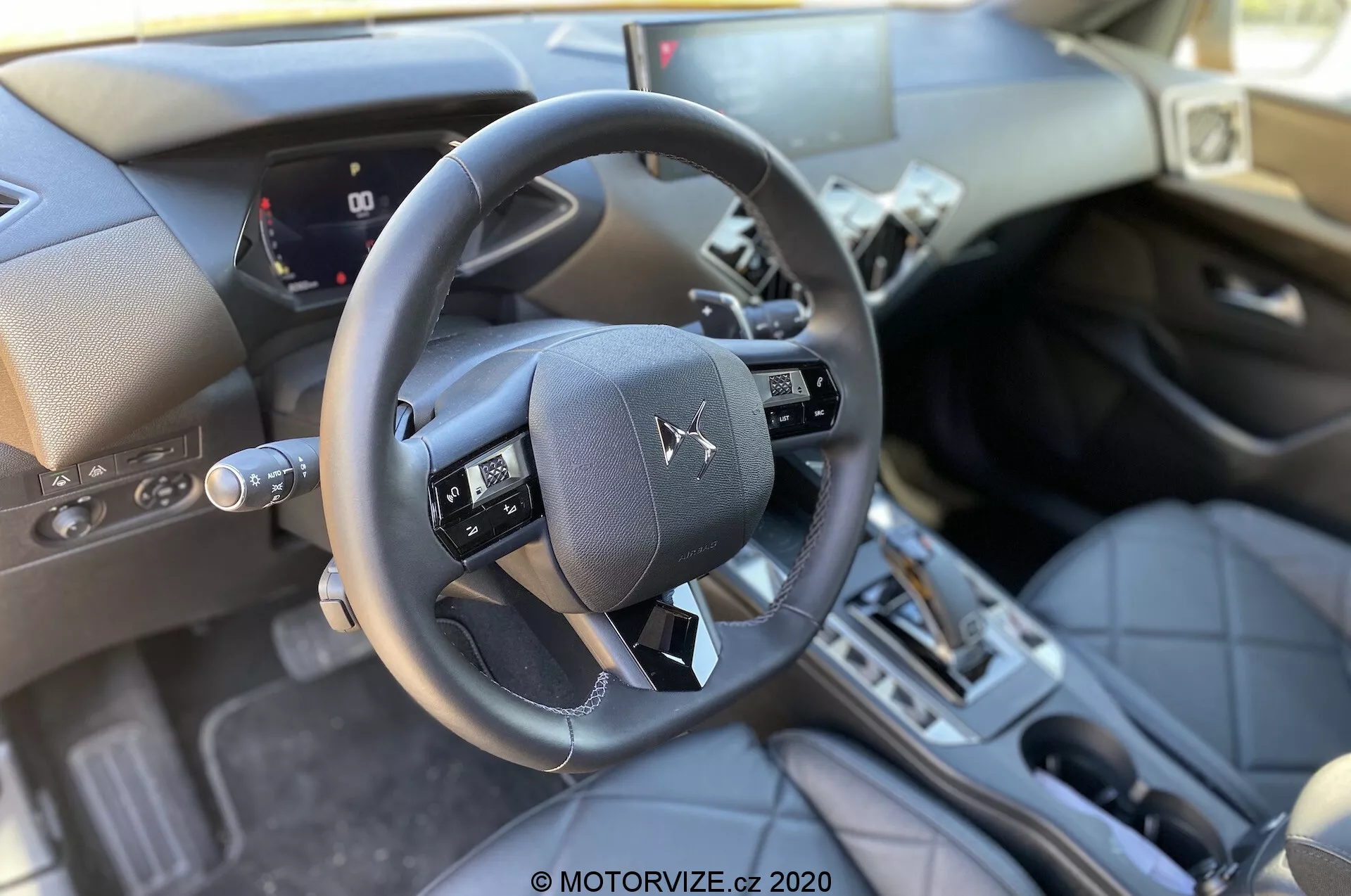 Vue intérieure de la DS Automobiles DS 3 Crossback 2019 depuis la perspective du siège passager avant, mettant en avant le volant à trois branches avec boutons multifonctions, tableau de bord numérique partiel affichant la vitesse de la voiture et les jauges, console centrale avec levier de changement de vitesse automatique, matériaux haut de gamme comme les surfaces en cuir et les accents métalliques, fente d'allumage traditionnelle, pédales et une lumière naturelle bien éclairée.
