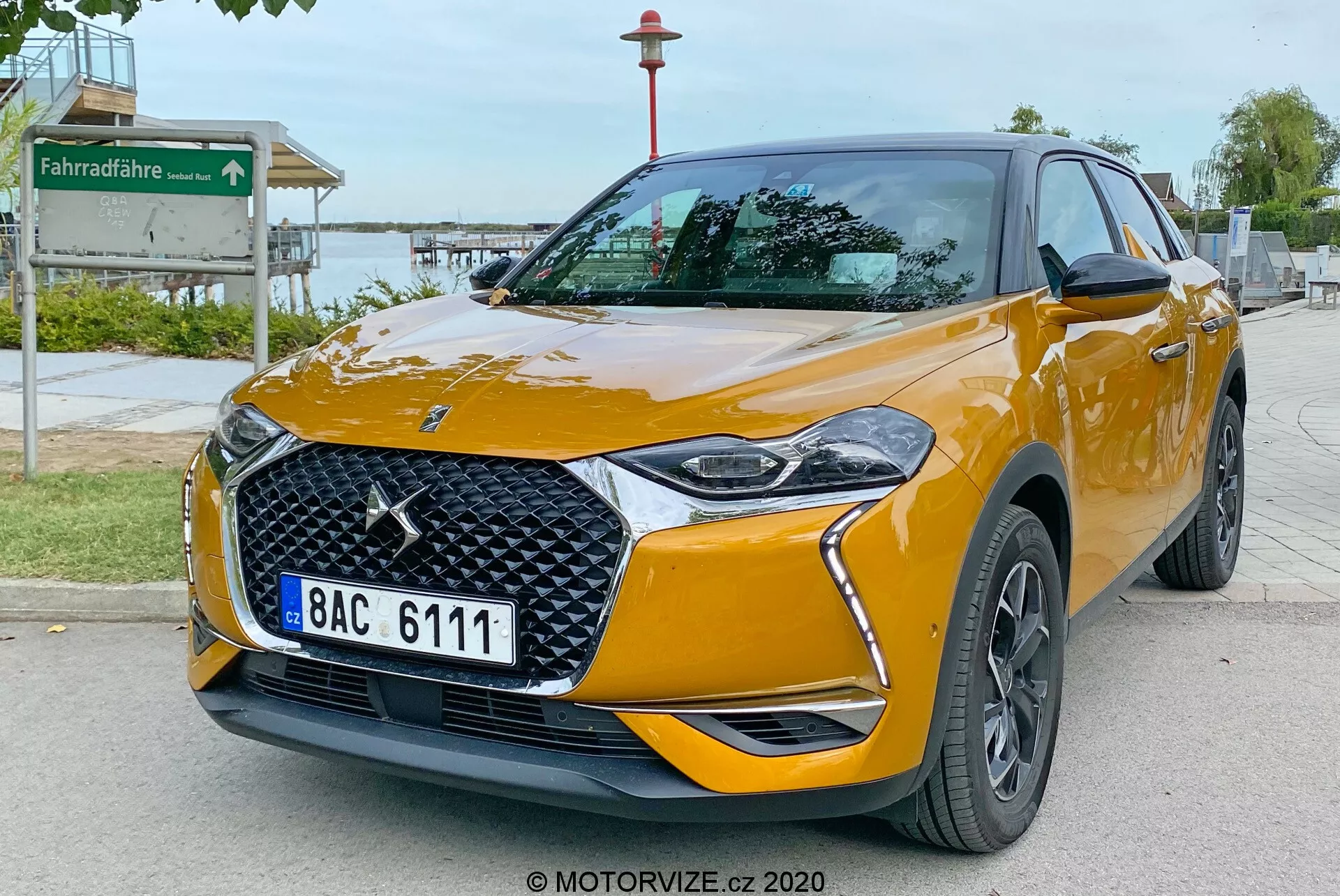 L'image montre un DS Automobiles DS 3 Crossback 2019, un SUV crossover compact moderne, stationné sur une zone pavée au bord de la route. Capturée sous un angle de trois quarts avant bas, l'image met en évidence la partie avant et le côté passager du véhicule. Les caractéristiques clés incluent un emblème de marque proéminent sur la grille à motif en losange, des phares LED élégants avec des feux de jour stylisés, des antibrouillards intégrés dans le pare-chocs avant, des rétroviseurs extérieurs couleur carrosserie avec clignotants intégrés et des roues en alliage à plusieurs branches de couleur sombre. La voiture est peinte en orange métallisé vibrant avec des lignes de carrosserie prononcées améliorant son aspect aérodynamique, et des badges pouvant indiquer les niveaux de finition. L'environnement suggère une zone publique près d'un parc, comme en témoigne un panneau de piste cyclable en arrière-plan.