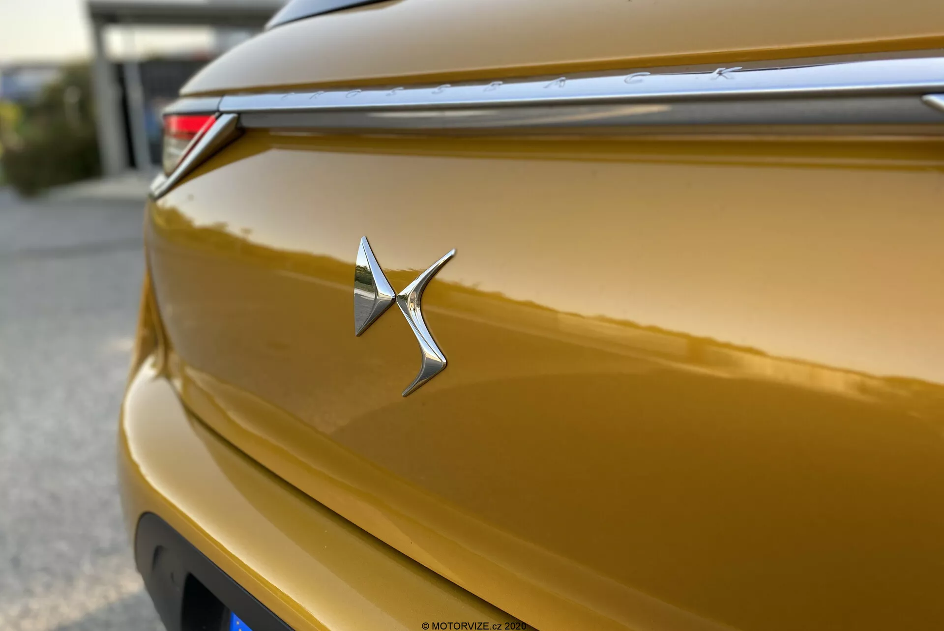 Gros plan d'une insigne du DS Automobiles DS-3 Crossback 2019 doré métallique. L'emblème chromé est net et placé au centre, ressemblant à une étoile avec un bras droit allongé. La carrosserie brillante reflète la lumière le long de ses courbes, montrant une riche teinte dorée. La faible profondeur de champ met en valeur l'insigne, avec une ligne horizontale au-dessus contribuant à son design aérodynamique. Un feu arrière à LED est partiellement visible dans le coin supérieur droit. Le couvercle du coffre arrière a une courbure subtile, et l'effet bokeh en arrière-plan isole l'insigne.
