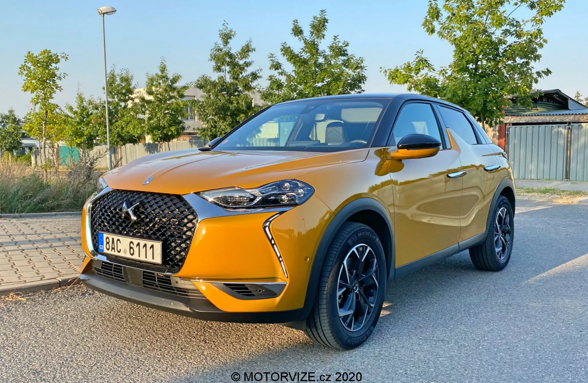 Vue trois quarts avant de la DS Automobiles DS 3 Crossback 2019, dévoilant la grande calandre distinctive à motif de grille et badge proéminent, phares LED élégants, peinture métallisée orange, jantes en alliage noir contrastantes, rétroviseurs latéraux intégrés et ligne des fenêtres ascendante se terminant par un spoiler aérodynamique. Le véhicule est garé avec du feuillage vert et des structures en arrière-plan flou, mettant en valeur le design et les caractéristiques sportives de la voiture.