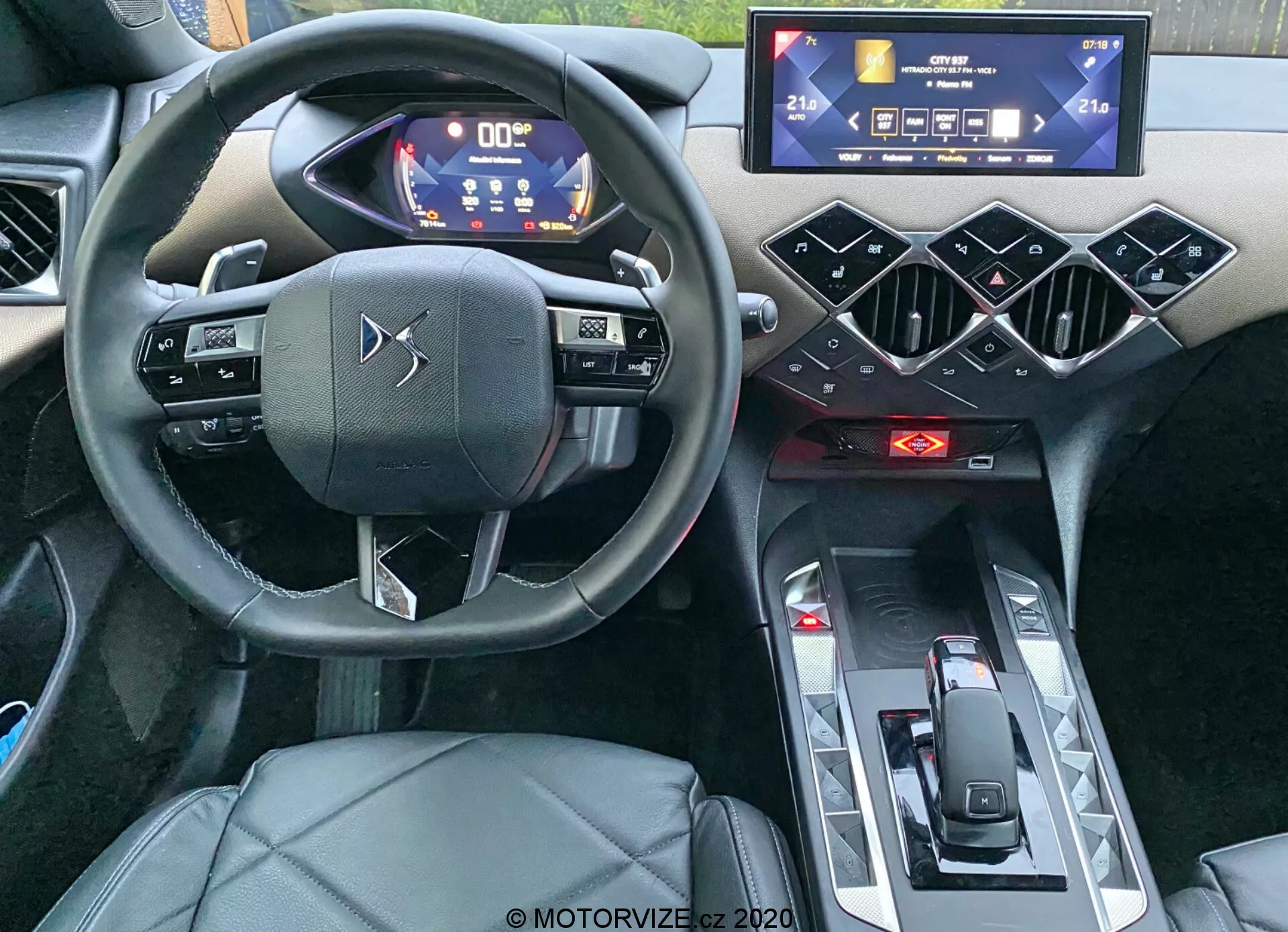 Vue intérieure de la DS Automobiles DS-3 Crossback 2019, se concentrant sur le poste de conduite depuis une perspective au niveau des yeux. La prise de vue, réalisée depuis l'arrière de l'habitacle, met en avant le volant à trois branches avec commandes tactiles capacitives, le tableau de bord numérique avec des compteurs éclairés en bleu et blanc, un système d'infodivertissement à écran tactile horizontal, des éléments de design hexagonaux géométriques sur la console centrale, des indicateurs de contrôle du climat rouges, un sélecteur de vitesse minimaliste et le siège conducteur en cuir ou similicuir. La photo souligne les caractéristiques modernes et high-tech ainsi que le design haut de gamme du véhicule.