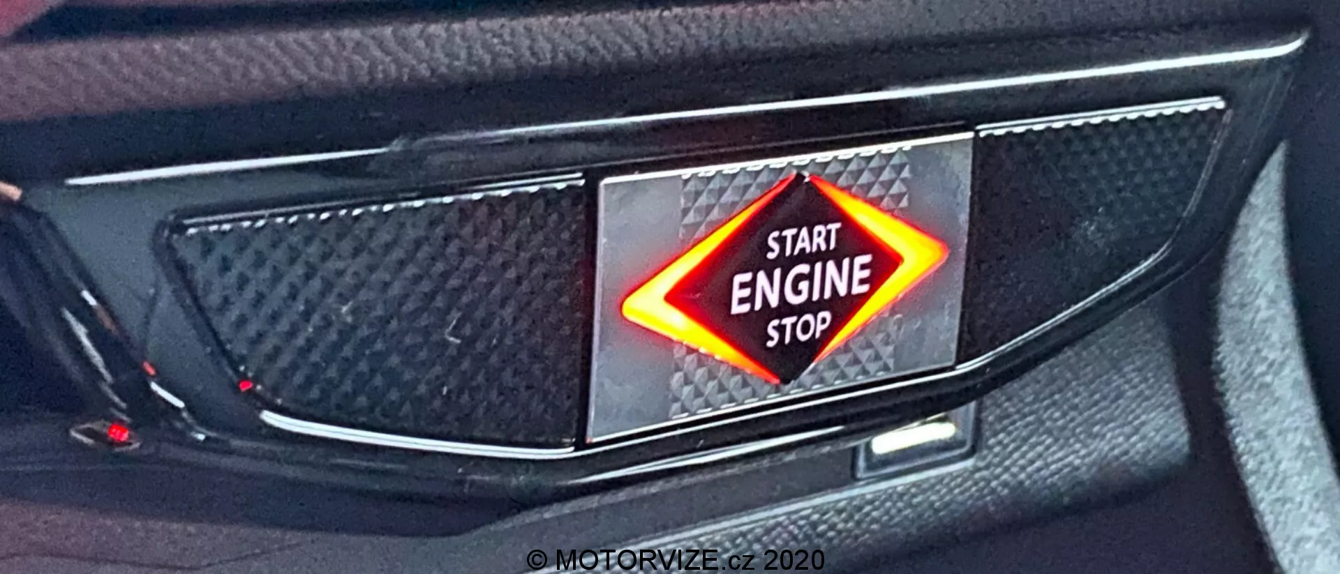 Gros plan sur le système de démarrage/arrêt du moteur à bouton-poussoir dans une DS Automobiles DS-3 2019. Le bouton hexagonal, situé sur la console centrale ou le tableau de bord, a une bordure illuminée rouge vif et un centre sombre, avec l'étiquette 'START ENGINE STOP' en caractères blancs gras. La texture en fibre de carbone du panneau environnant, combinée avec des garnitures noires brillantes, met en avant le design intérieur sportif et luxueux du véhicule.