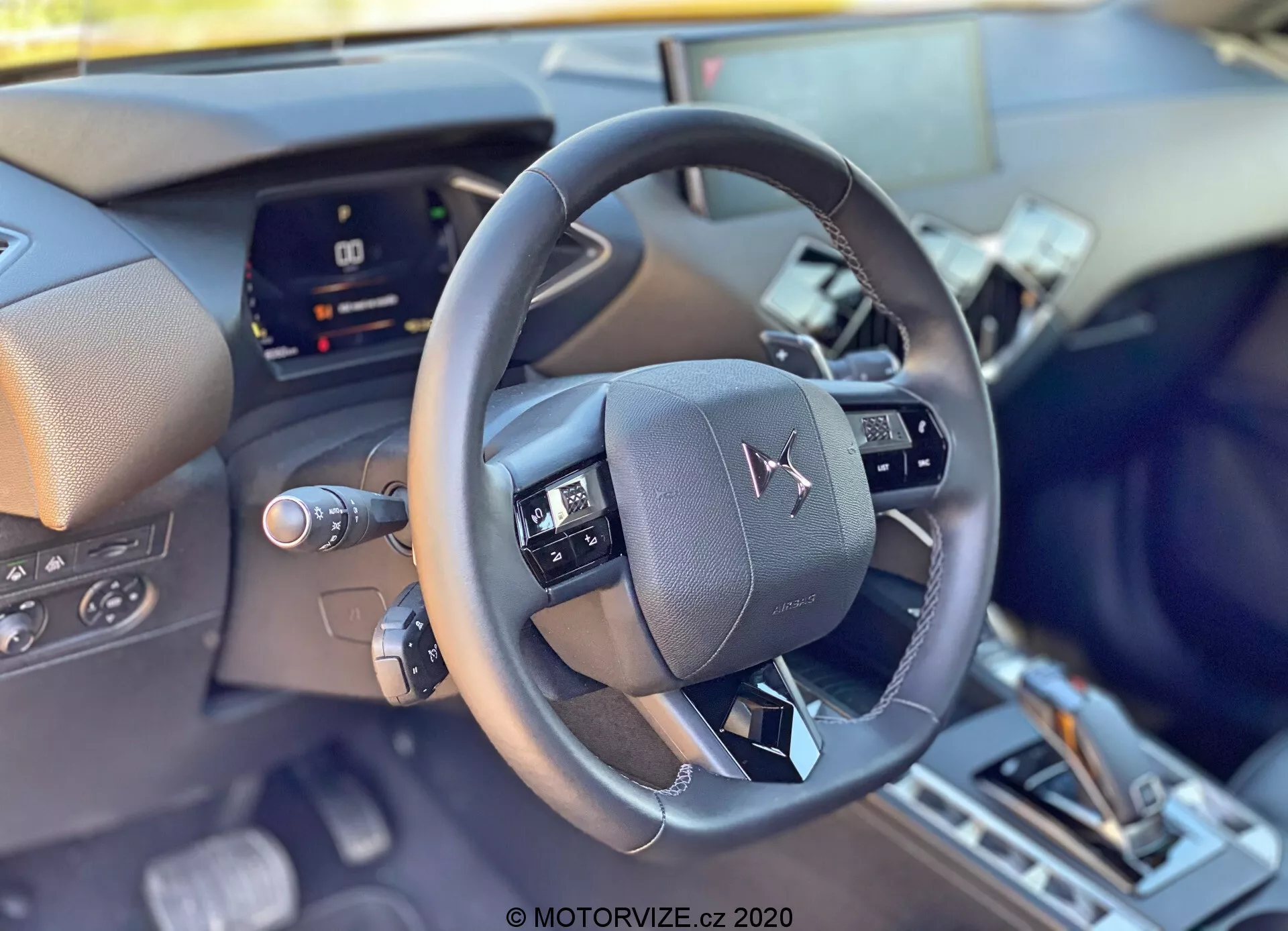 Vue intérieure en gros plan de la DS Automobiles DS-3 2019 (DS-3 Crossback) depuis la perspective du conducteur, centrée sur le volant en cuir moderne avec des commandes multifonctions. Derrière le volant, on trouve un tableau de bord numérique partiellement obstrué, indiquant une technologie avancée du véhicule. La partie inférieure laisse entrevoir la console centrale avec un levier de vitesses et d'éventuels sélecteurs de modes de conduite, reflétant des caractéristiques intérieures et de confort sophistiquées.