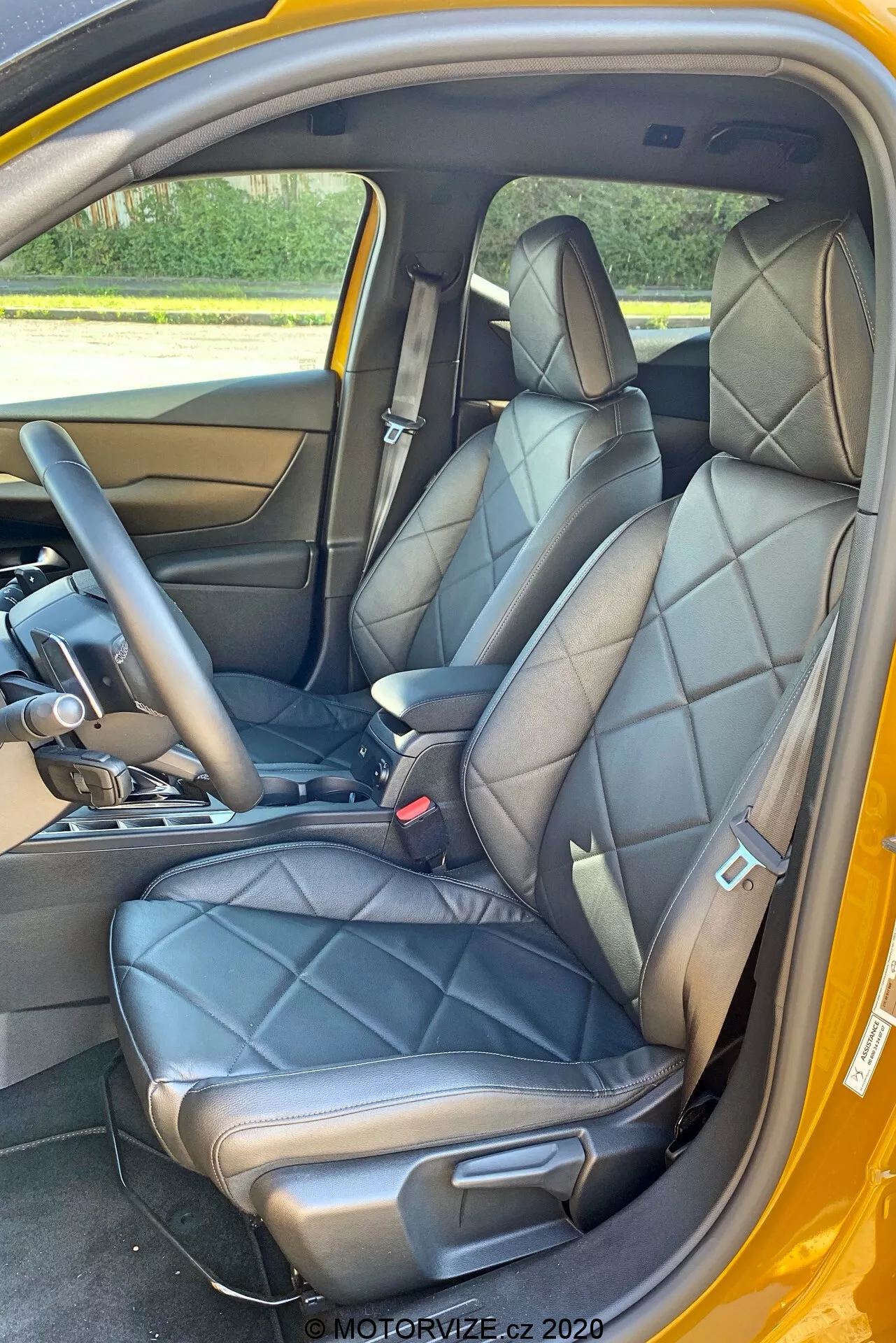 Vue intérieure d'une DS Automobiles DS-3 Crossback 2019 se concentrant sur les sièges avant depuis la porte latérale du passager. L'image met en lumière des sièges en cuir noir matelassé avec des appuie-têtes intégrés et des supports latéraux, suggérant un design luxueux ou sportif. Une partie de la console centrale avec un levier de vitesses indiquant une transmission automatique est visible, ainsi que la partie inférieure du tableau de bord. Le panneau intérieur de la porte correspond au cuir des sièges, avec une poignée de porte en métal et des boutons de commande. On voit également des ceintures de sécurité noires avec un mécanisme de réglage en hauteur, un plancher en moquette et un schéma de couleur extérieure contrastant, possiblement jaune.