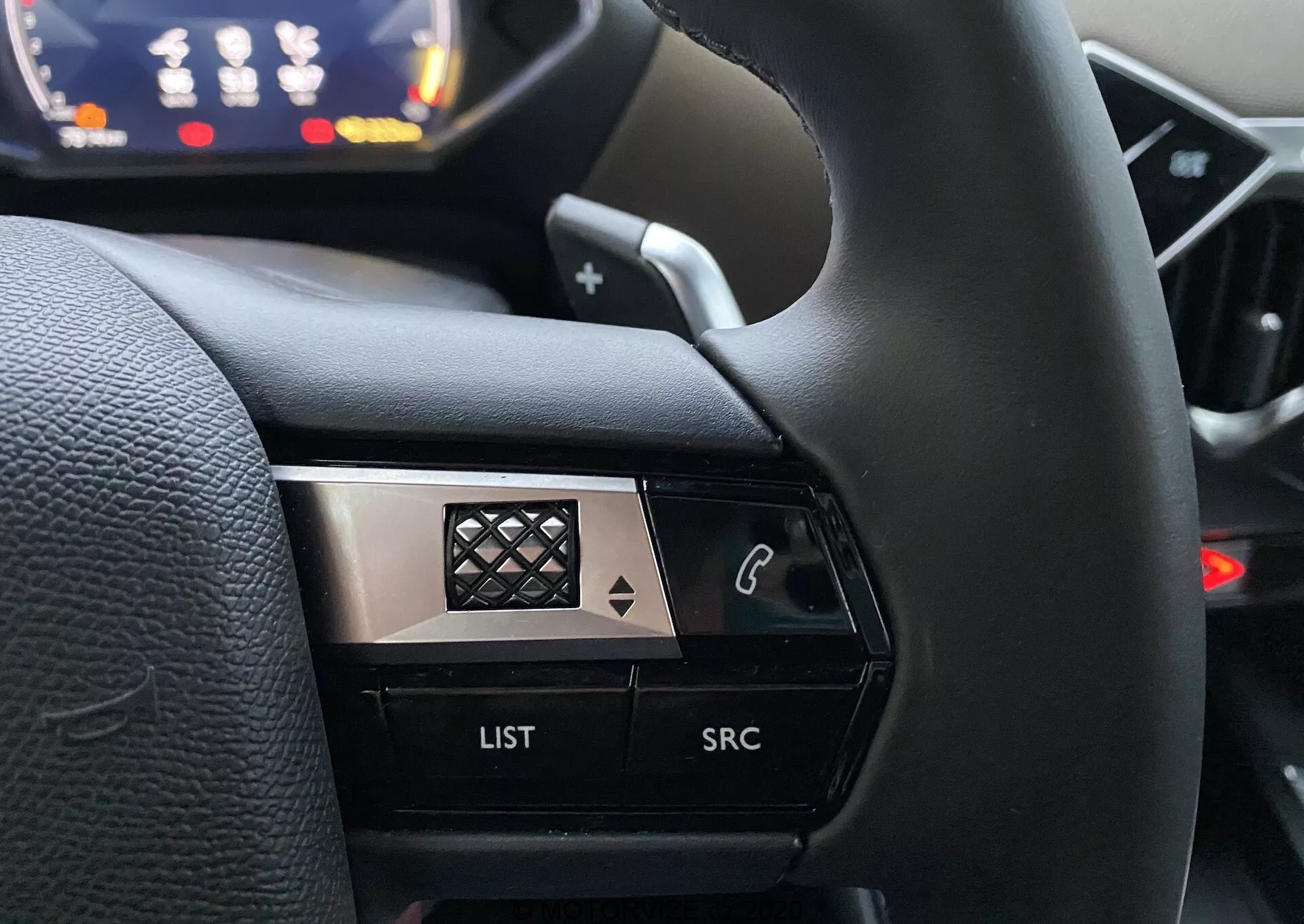 Vue rapprochée depuis la perspective du conducteur du côté gauche d'un volant dans une DS Automobiles DS 3 Crossback 2019. L'image met en avant les boutons de contrôle, y compris un bouton avec icône de téléphone, un bouton 'LIST' et un bouton 'SRC', mis en valeur devant un tableau de bord flou. Le design à fort contraste combine des finitions noires et métalliques avec un motif en diamant sur le chrome, rehaussant son aspect premium.