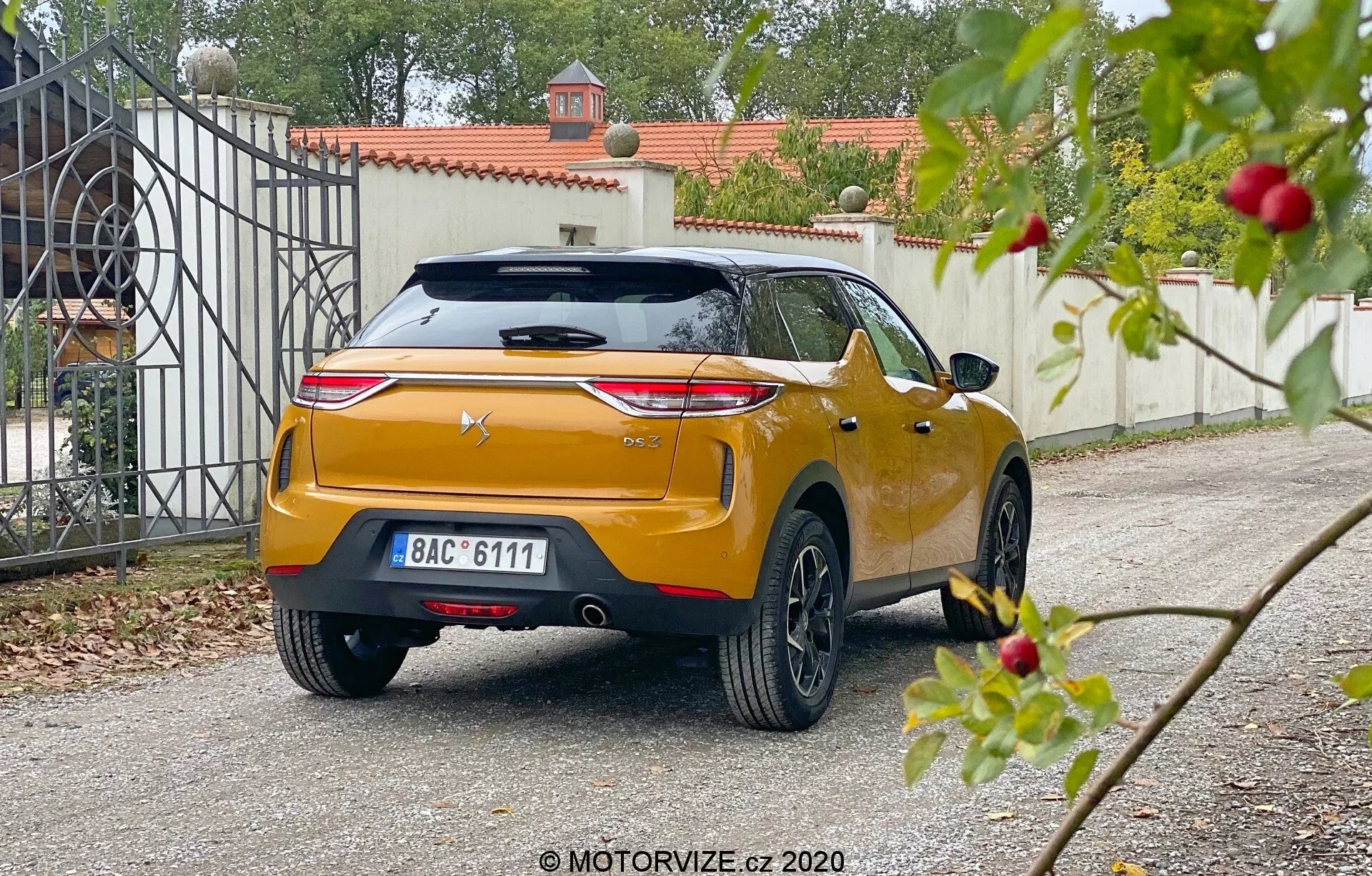 Vue arrière en angle d'une DS Automobiles DS 3 Crossback 2019, capturée sous une perspective basse dans une allée résidentielle. La voiture arbore des feux arrière horizontaux proéminents, un logo du fabricant centré sur un hayon sculpté avec un spoiler intégré et un troisième feu stop, une plaque d'immatriculation centrée et une finition métallique brillante de couleur orange ou dorée. Le pare-chocs arrière massif a des capteurs ou des réflecteurs intégrés, avec essuie-glace arrière visible, antenne à aileron de requin et roues en alliage à rayons multiples. Le feuillage légèrement flou au premier plan contraste avec la mise au point plus nette sur la voiture et la porte d'entrée à l'arrière-plan, suggérant une lumière du jour douce et diffuse.