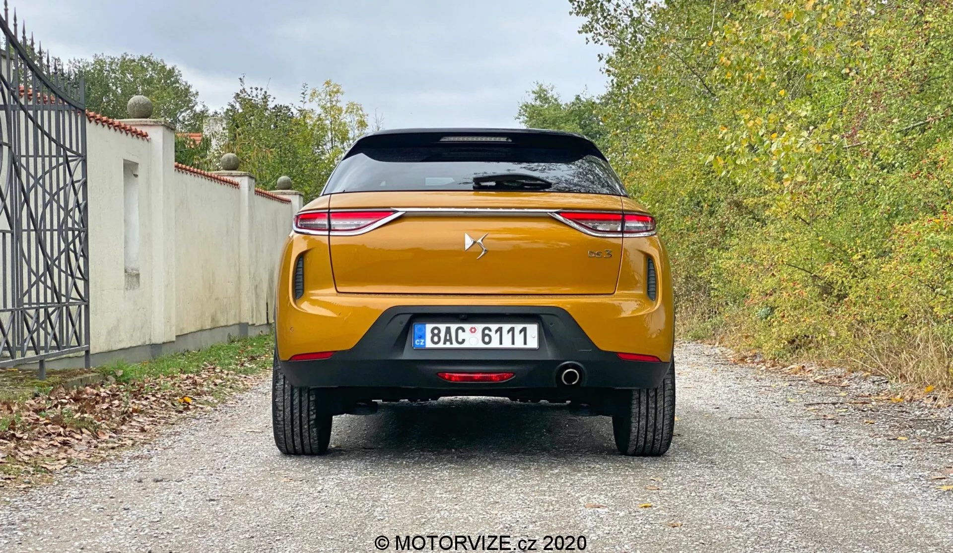 Vue arrière d'une voiture moderne DS-Automobiles DS-3 Crossback 2019 montrant ses caractéristiques de design et de technologie, y compris un aileron monté sur le toit, un pare-brise arrière avec élément chauffant, un feu stop en hauteur, un couvercle de coffre avec logo du fabricant, une bande chromée proéminente, des feux arrière LED élégants, un pare-chocs arrière robuste avec des capteurs de stationnement, un système d'échappement double, un élément diffuseur noir et une couleur orange ou cuivre frappante sur une surface de gravier avec végétation en arrière-plan.