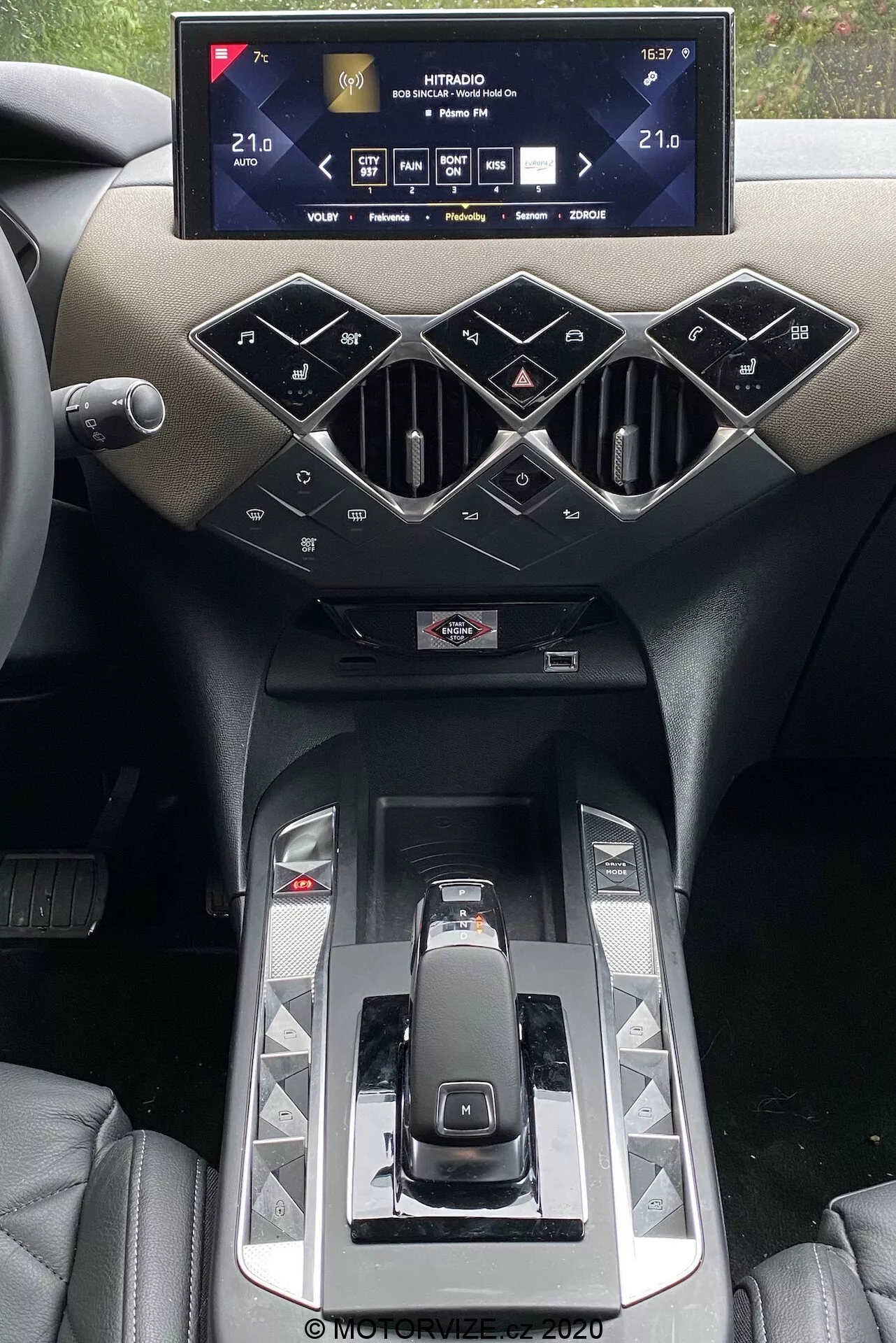 Intérieur de la DS Automobiles DS 3 2019 mettant en avant la console centrale avec un système d'info-divertissement. Un large écran tactile horizontal affiche les réglages du véhicule, y compris les températures de contrôle du climat réglées à 21,0°C. En dessous, des bouches d'aération au style géométrique et des boutons physiques en disposition en diamant contrôlent les systèmes de la voiture. Le levier de vitesses moderne, les panneaux brillants et un aperçu de portes-gobelets sont mis en valeur par des matériaux luxueux et un design haut de gamme.