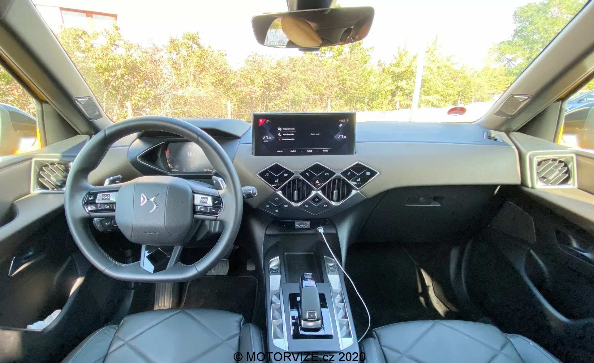 Intérieur d'une DS Automobiles DS-3 2019 (DS-3 Crossback) moderne vu de la perspective de la banquette arrière, montrant la zone de la cabine avant avec tableau de bord, volant, console centrale avec levier de vitesses, porte-gobelets, panneau de contrôle du climat tactile, écran tactile d'infodivertissement proéminent, volant multifonction, bouches d'aération angulaires, sièges avant en cuir avec surpiqûres contrastantes, garniture de porte avec boutons de commande et toit panoramique.