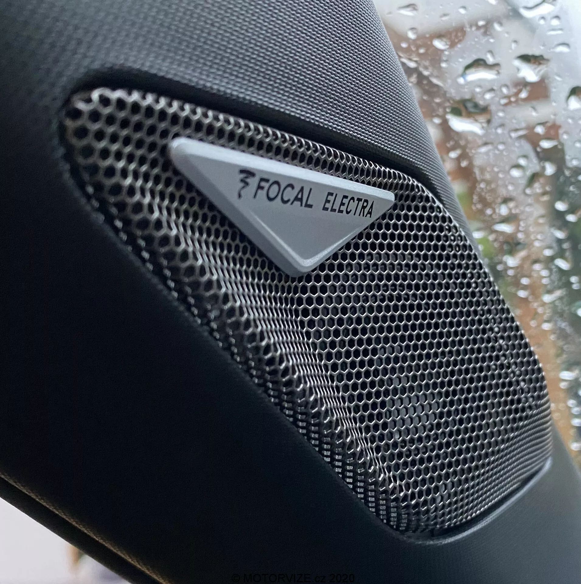 Gros plan sur le haut-parleur de marque Focal Electra dans la DS Automobiles DS-3 Crossback 2019, mettant en avant le composant audio haute-fidélité avec une grille en nid d'abeille en métal de haute qualité et un logo Focal et une marque Electra clairs. La prise de vue se concentre sur le design complexe du haut-parleur, avec une profondeur de champ réduite qui met en valeur la texture du haut-parleur contre un arrière-plan flou de la fenêtre de la voiture avec des gouttes de pluie.