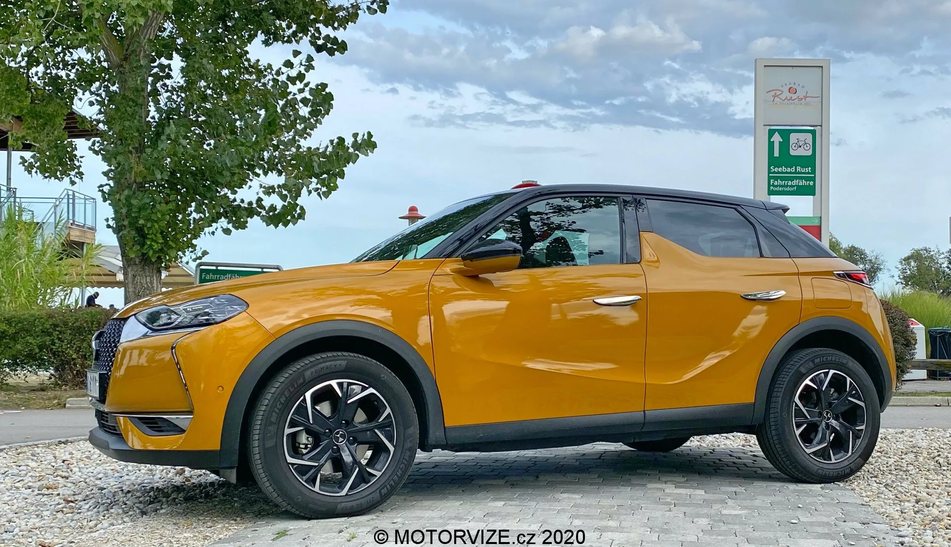 Profil latéral d'un DS Automobiles DS 3 Crossback 2019 orange vif, stationné près d'un bord de route avec verdure et panneau de station-service. Le côté conducteur, avec des roues noires à rayons multiples et des rétroviseurs latéraux de la couleur de la carrosserie, est visible sous un angle bas à hauteur des roues de la voiture, montrant son design élégant de SUV compact avec une ligne de ceinture ascendante et des vitres teintées.