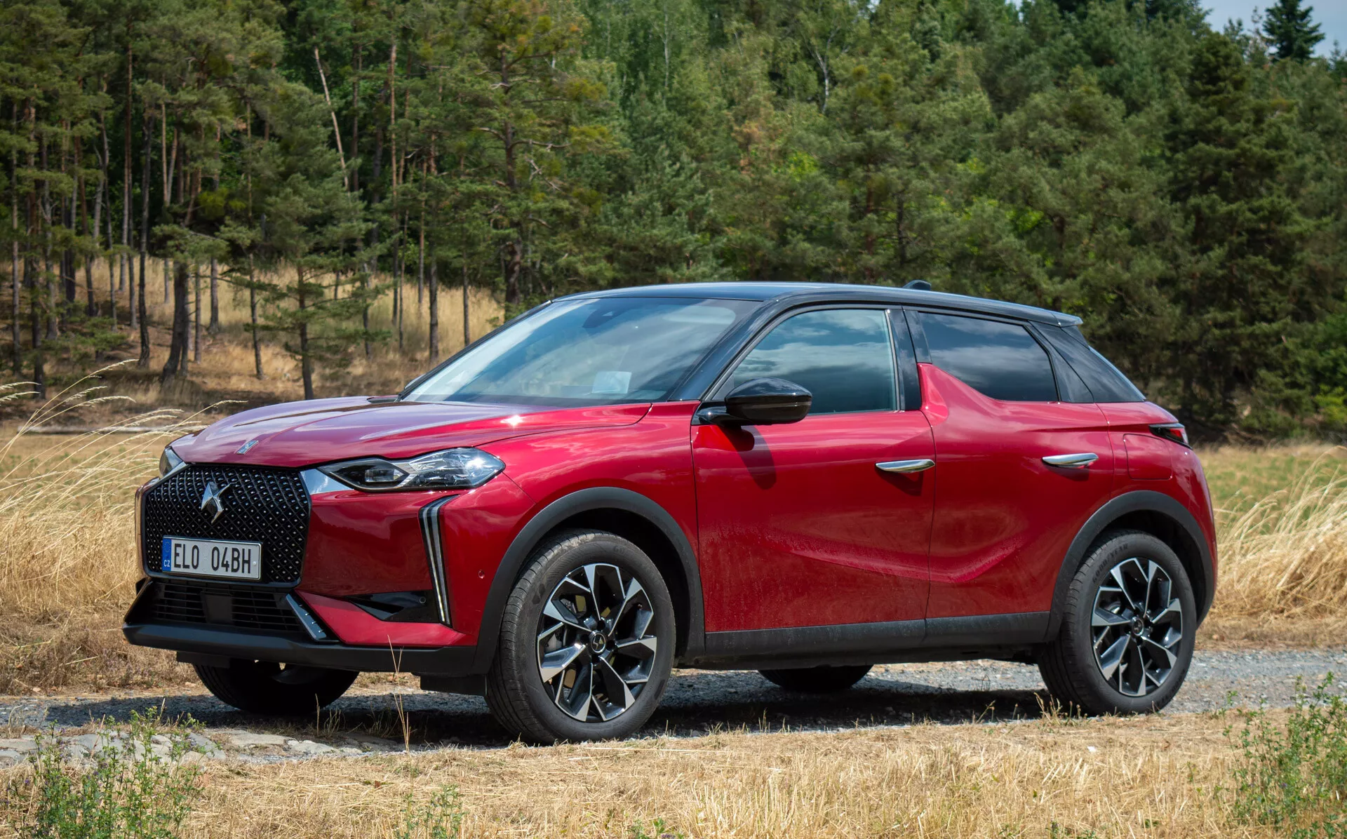 Vue de côté avant de la DS Automobiles DS 3 E-TENSE 2023, SUV crossover rouge, jantes en alliage à multiples branches, phares LED, sur un chemin de gravier avec un arrière-plan forestier