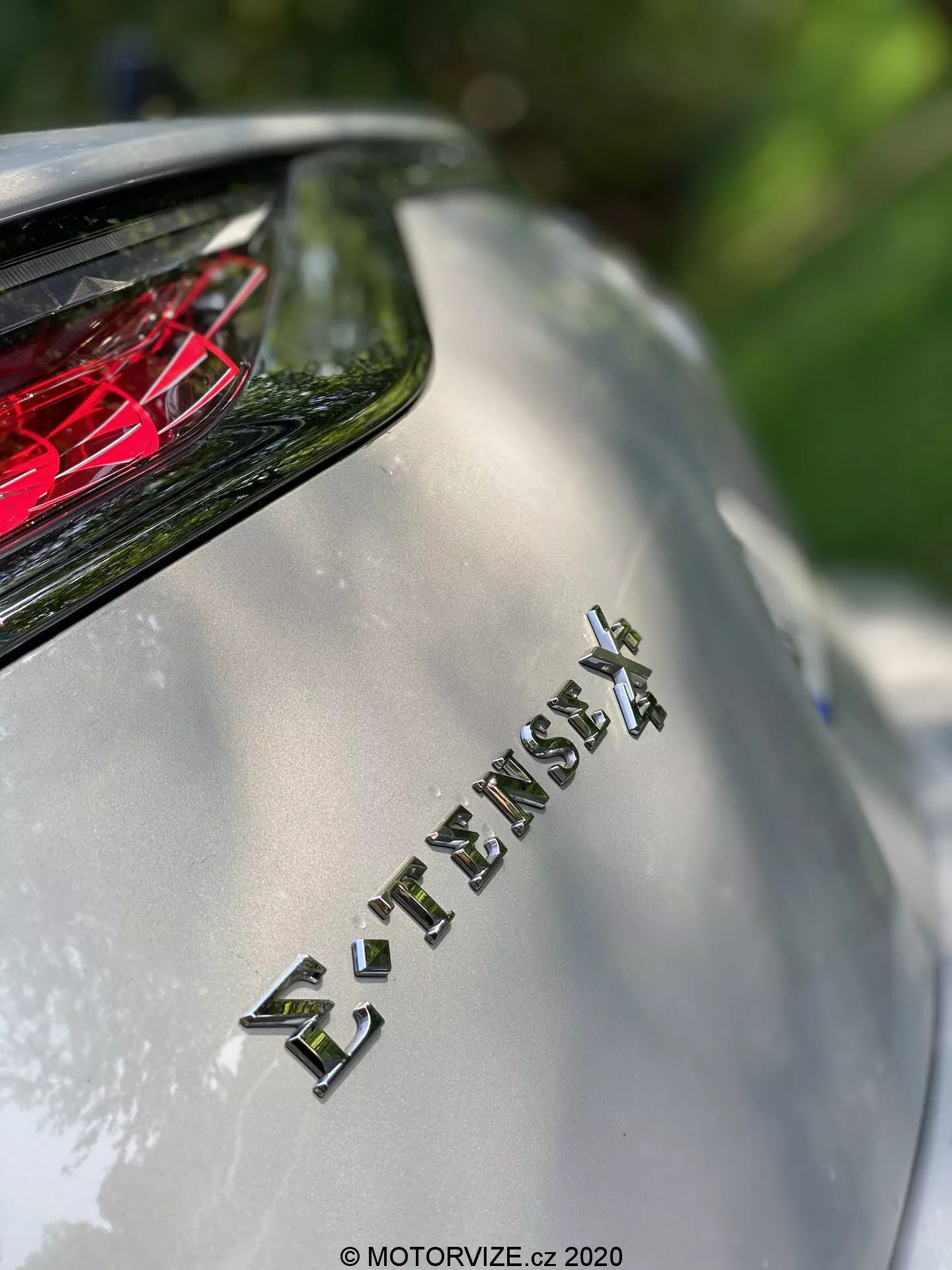Gros plan sur la section arrière du modèle DS Automobiles DS 7 Crossback 2018, se concentrant sur l'emblème 'E-TENSE 4X4' indiquant la transmission intégrale et probablement une motorisation hybride ou électrique. La faible profondeur de champ floute l'arrière-plan tout en mettant en avant l'emblème, avec le feu arrière cristallin rouge visible au-dessus. L'image capture la finition métallique de la carrosserie et le lettrage chromé sous la lumière naturelle du soleil, soulignant la technologie avancée et le design luxueux du véhicule.