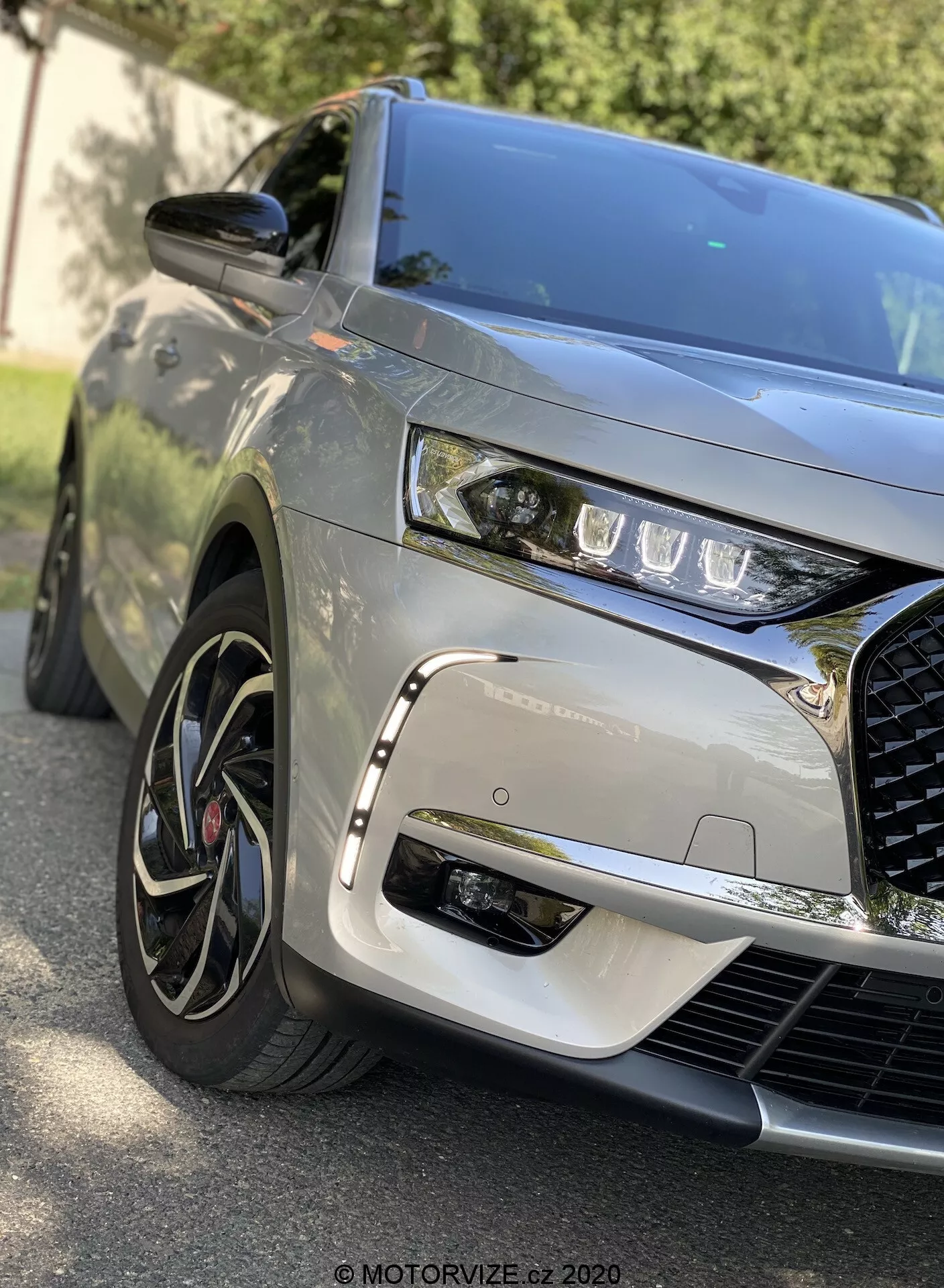 Plan rapproché avec angle de vue avant du DS Automobiles DS 7 Crossback 2018 capturant le quart avant gauche, montrant des phares LED élégants, une grille proéminente et complexe, un capot sculpté, des roues en alliage à plusieurs rayons avec pneus à profil bas, une finition de peinture métallique, un rétroviseur latéral de la couleur de la carrosserie et des reflets dynamiques indiquant un environnement extérieur.