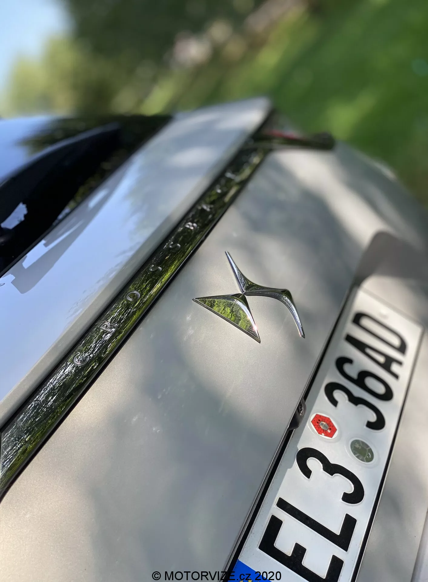 Photo en gros plan anglé de la partie arrière d'une DS Automobiles DS 7 Crossback argentée de 2018. L'image met en avant l'emblème métallique de la voiture centré sur le couvercle du coffre arrière, une plaque d'immatriculation de style européen avec des caractères noirs 'EL3 36AD' sous l'emblème, et le bord supérieur du pare-chocs arrière. Une partie du pare-brise arrière avec le dégivreur visible, ainsi que des surfaces réfléchissantes à haute brillance, apparaît également. La faible profondeur de champ met en évidence l'emblème tout en floutant doucement les bords du couvercle du coffre et du pare-brise.