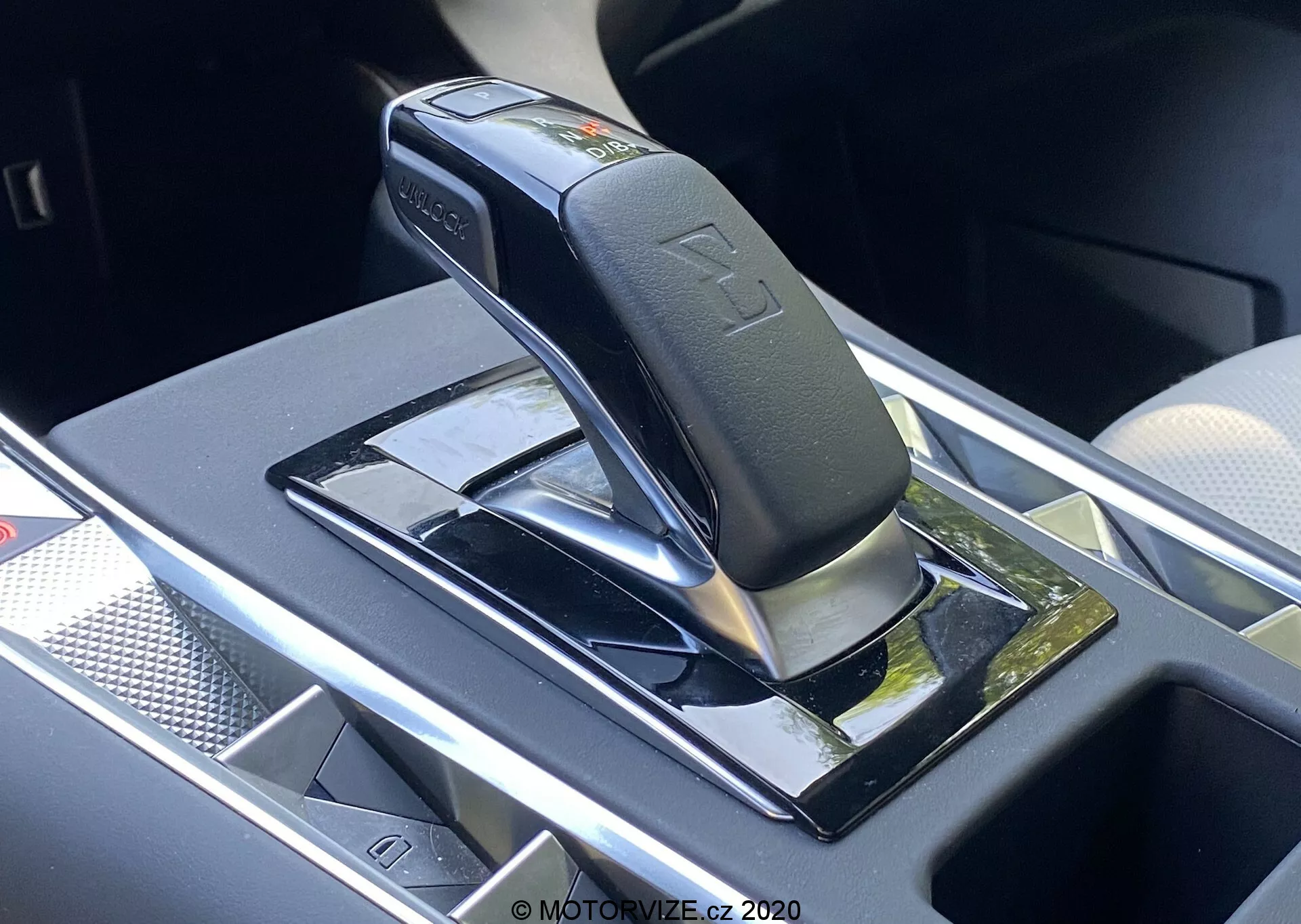 Vue en gros plan du levier de changement de vitesses automatique de la DS Automobiles DS-7 2018 (DS-7 Crossback), montrant un levier noir brillant avec des accents argentés, probablement en position de parking ou neutre, monté sur un panneau noir piano avec des surfaces réfléchissantes. Les caractéristiques incluent des symboles en relief pour les positions des vitesses, un bouton de sécurité sur le levier et un compartiment de rangement à proximité avec un éventuel chargeur sans fil. La zone environnante présente des garnitures chromées ou argentées, indiquant un intérieur de véhicule moderne et luxueux.