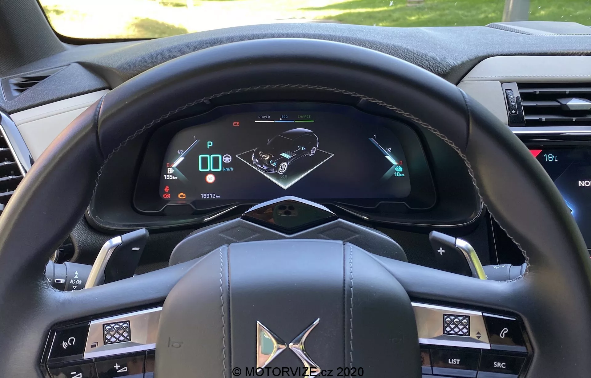 Vue rapprochée du côté conducteur d'une voiture moderne depuis la perspective de l'occupant montrant un volant multifonction en cuir avec des accents métalliques et divers boutons de commande, un tableau de bord entièrement numérique affichant la vitesse, la vitesse engagée et une carte de navigation, des leviers de commande des clignotants et des essuie-glaces sur les branches du volant, des boutons pour le système d'infotainment et de climatisation sur le tableau de bord, des palettes de changement de vitesse semi-automatiques, un bouton start/stop, et des matériaux intérieurs de haute qualité.