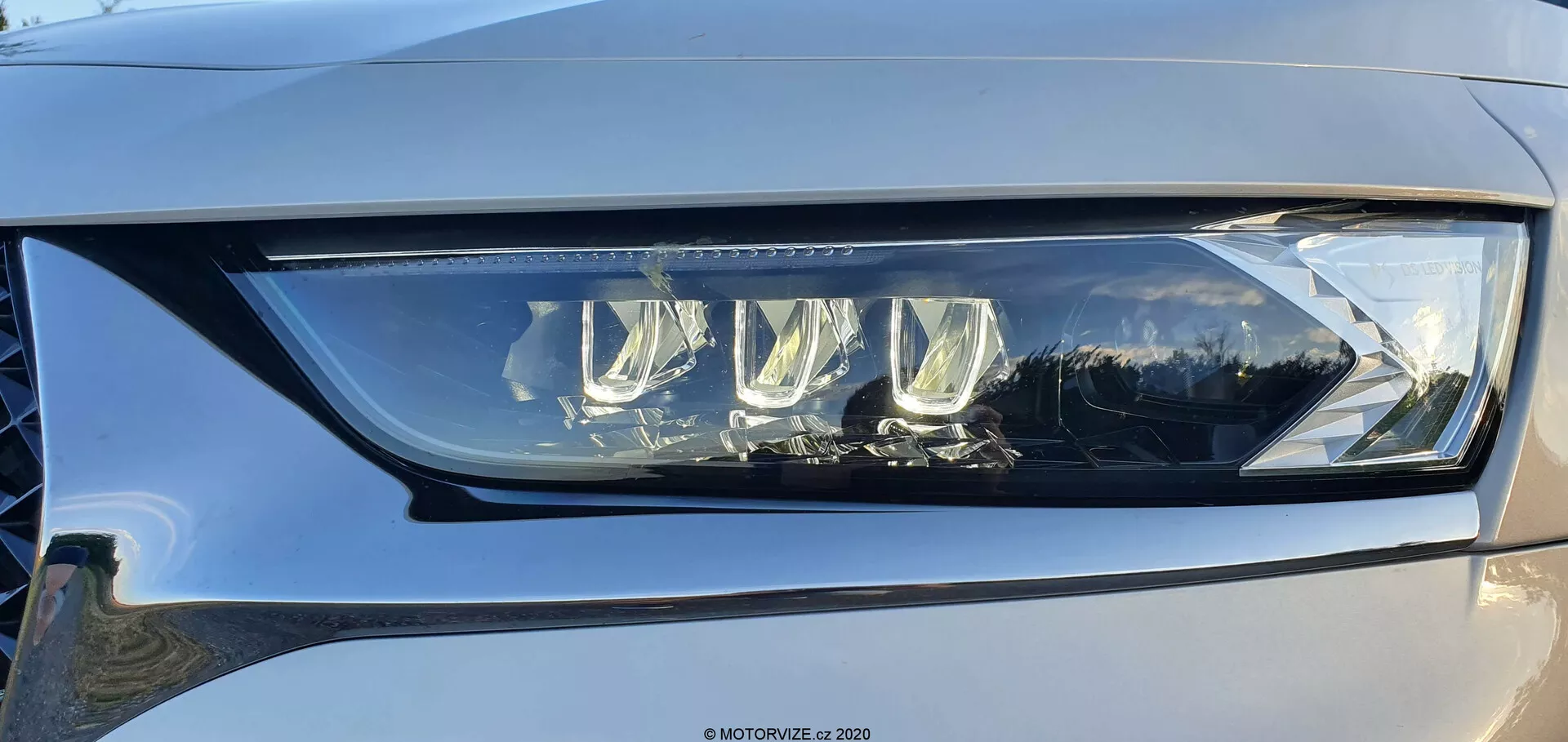 Gros plan de l'assemblage du phare gauche du DS Automobiles DS-7 Crossback sous un angle léger, montrant un design LED moderne avec plusieurs lentilles de projecteur et une distinctive lumière de jour LED, le tout encapsulé dans un plastique transparent de haute qualité. Le phare illustre une technologie d'éclairage automobile avancée capable de fournir des fonctions de faisceaux bas et haut, avec des caractéristiques adaptatives potentielles pour améliorer la visibilité. Un léger reflet sur la surface brillante et une touche de couleur gris clair à moyen de la voiture peuvent également être vus.