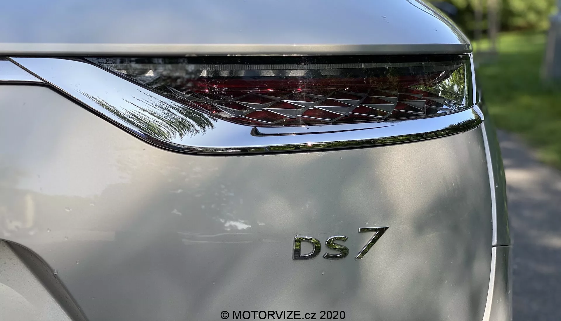 Vue en gros plan de la section arrière d'une DS Automobiles DS 7 Crossback 2018 capturée d'une perspective en contre-plongée. L'accent est mis sur le hayon, mettant en évidence les lettres en gras 'DS' au-dessus de la désignation du modèle '7'. Au-dessus du nom du modèle se trouve un feu arrière incurvé et étendu horizontalement avec des éléments LED distinctifs. La couleur de la carrosserie est argent métallisé et le design sculpté suggère une apparence moderne et luxueuse. La photographie met en avant le branding et le design du feu arrière, avec un environnement extérieur lumineux subtilement reflété sur la surface de la voiture.