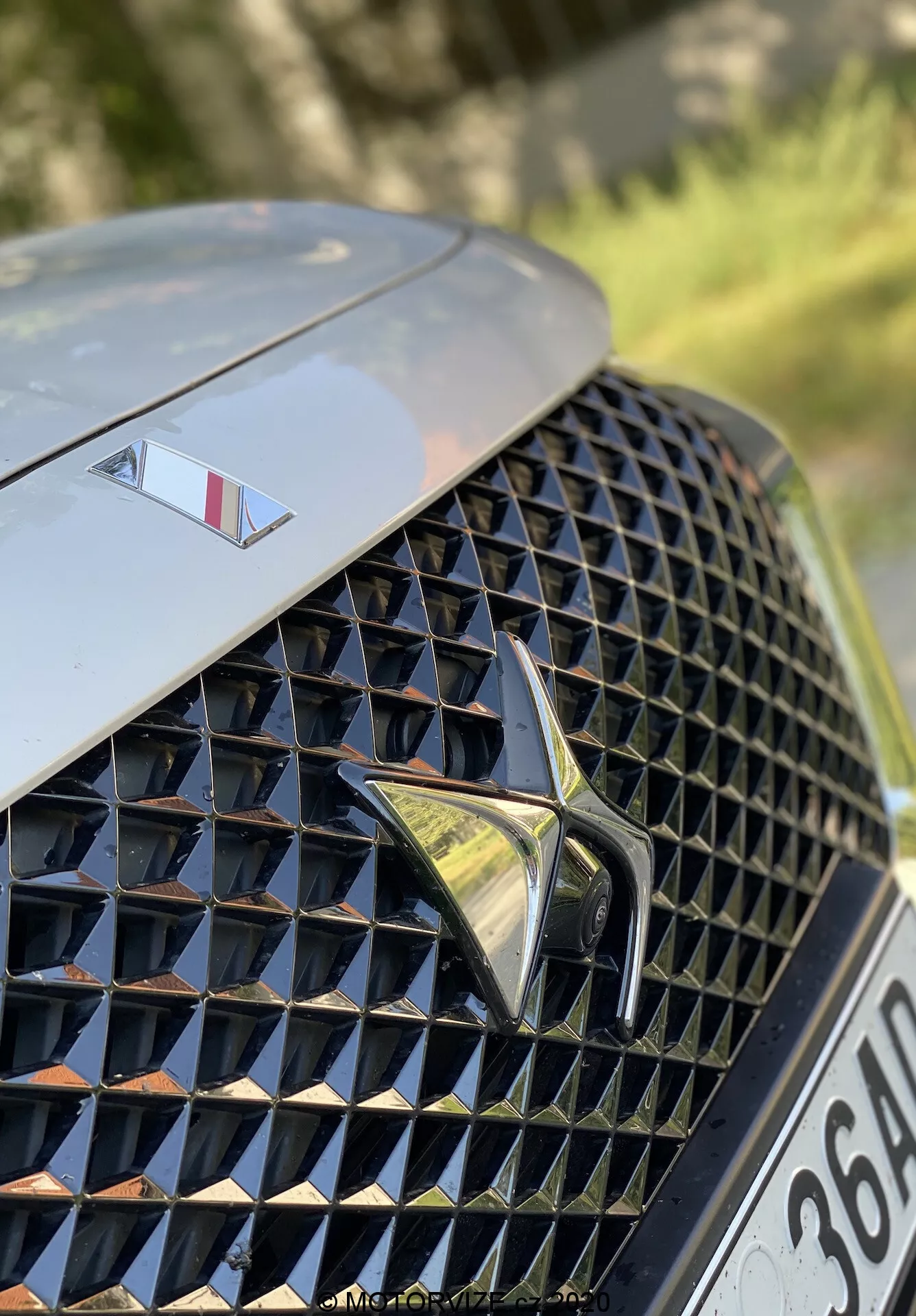 Gros plan sur la section avant gauche du DS Automobiles DS 7 Crossback 2018. L'image se concentre sur la grille avant luxueuse à motif en diamant complexe avec des surfaces réfléchissantes, centrée avec un logo stylisé proéminent. Au-dessus de la grille, le capot présente un plus petit emblème signifiant un modèle haut de gamme. L'arrière-plan est flou, mettant en valeur le design de la grille et les éléments de marque.