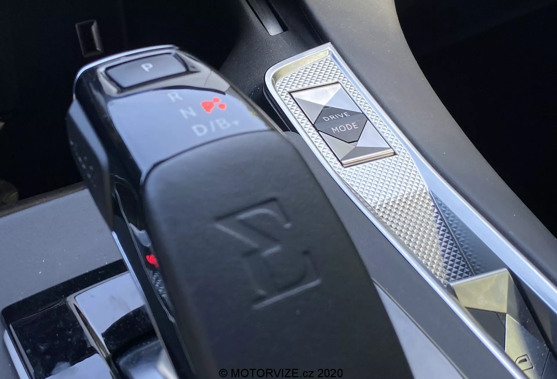Vue rapprochée de la console centrale d'une DS 7 Crossback 2018 mettant en avant le sélecteur de vitesses et le sélecteur de modes de conduite depuis un angle oblique. Le sélecteur de vitesses électronique en forme de L indique la position Park avec une lumière LED rouge et une fonction de bouton. À droite, un sélecteur de modes de conduite avec motif diamant et une garniture argentée indiquent la finition intérieure haut de gamme et les systèmes avancés de contrôle du véhicule.