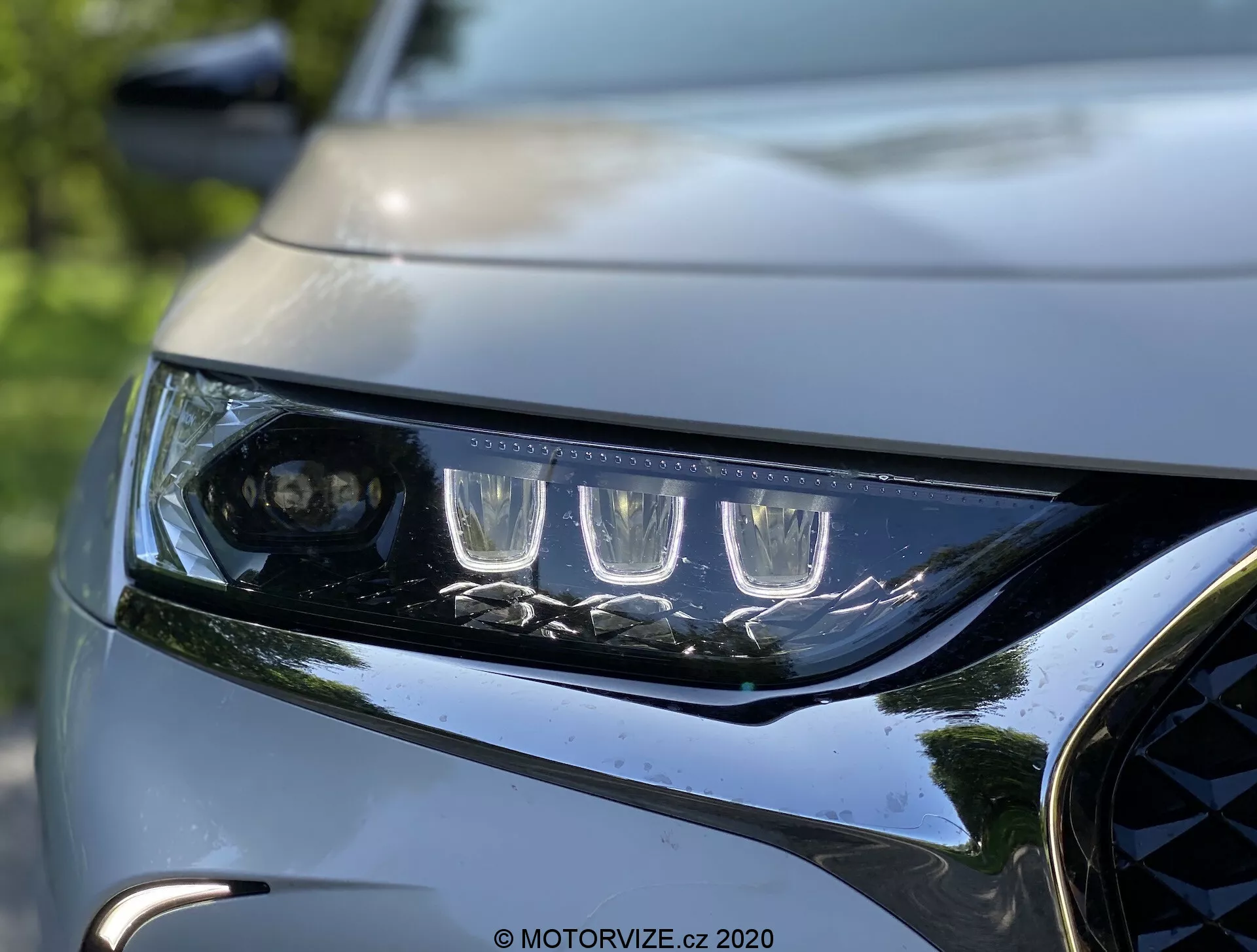 Vue rapprochée du phare gauche et de la calandre avant du DS Automobiles DS 7 Crossback 2018. Le phare comporte des feux de jour à LED distinctifs et une lentille de projecteur pour le faisceau principal, encadré par plusieurs éléments LED. Le boîtier de phare élégant et angulaire montre l'aérodynamisme moderne. Une partie de la calandre sombre et brillante avec des accents métalliques et une partie du capot sculpté mettent en valeur le design dynamique et premium du véhicule. L'arrière-plan est flou, soulignant la netteté et les détails complexes de l'éclairage et de la face avant de la voiture.