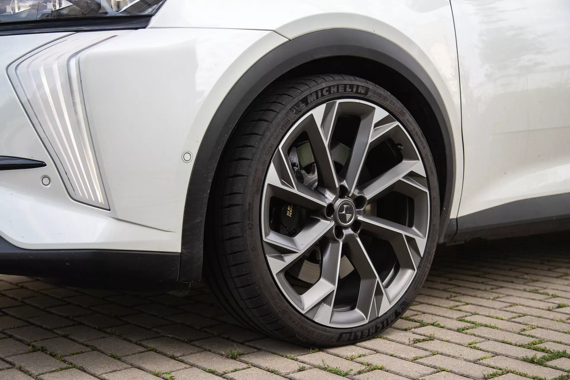 Vue rapprochée de l'ensemble roue avant d'un DS-Automobiles DS-7 2022 sur surface en briques imbriquées, mettant en avant une roue en alliage métallique à multiples branches avec capuchon central logo, pneu radial Michelin avec motifs de bande de roulement détaillés, aile avant blanche au design angulaire et garde-boue noir, sous un éclairage naturel