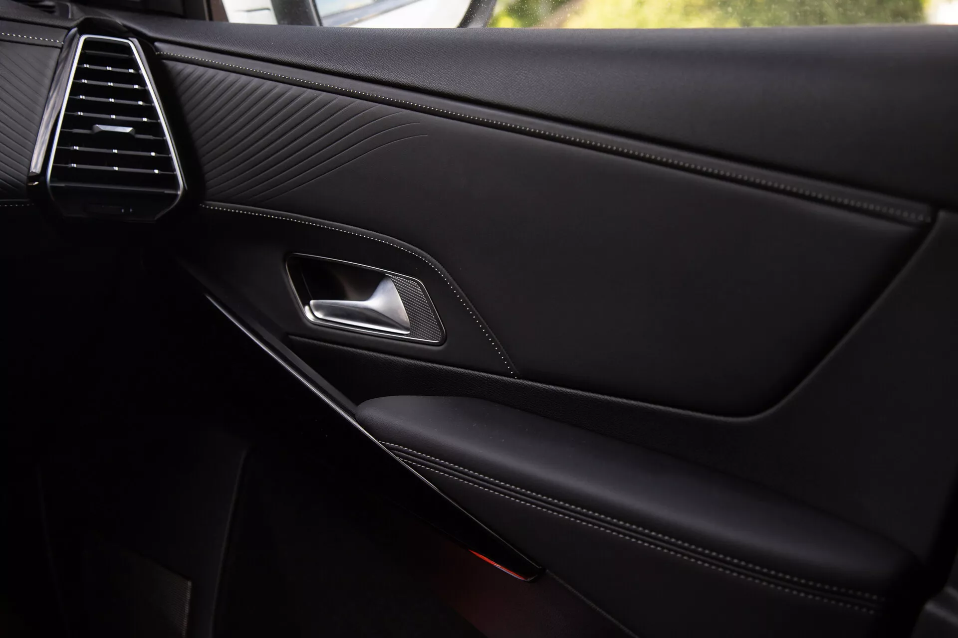 Vue rapprochée du panneau de porte intérieur de la DS Automobiles DS-7 2022 mettant en évidence la grille d'aération élégante, la texture en cuir piqué, la poignée de porte en chrome, le levier de verrouillage ergonomique, l'accoudoir à coutures contrastées et le compartiment de rangement intégré avec une grille de haut-parleur partiellement visible, reflétant une qualité premium et des détails de conception.