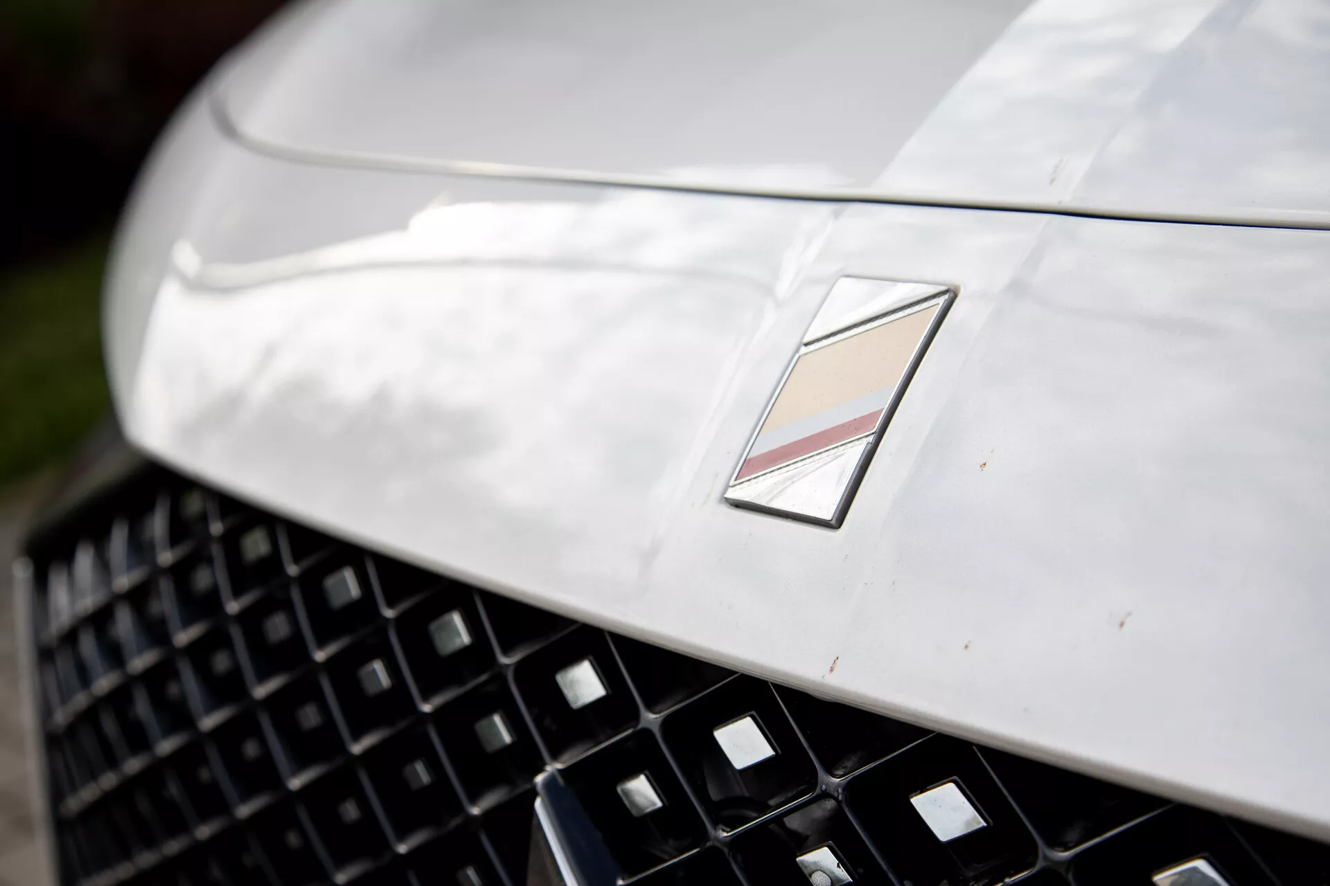 Vue rapprochée de la section avant d'une voiture de luxe montrant un capot métallique au design aérodynamique, une grille détaillée pour une meilleure admission d'air, et un emblème multifacette signifiant le fabricant, sous réflexion de lumière naturelle et faible profondeur de champ