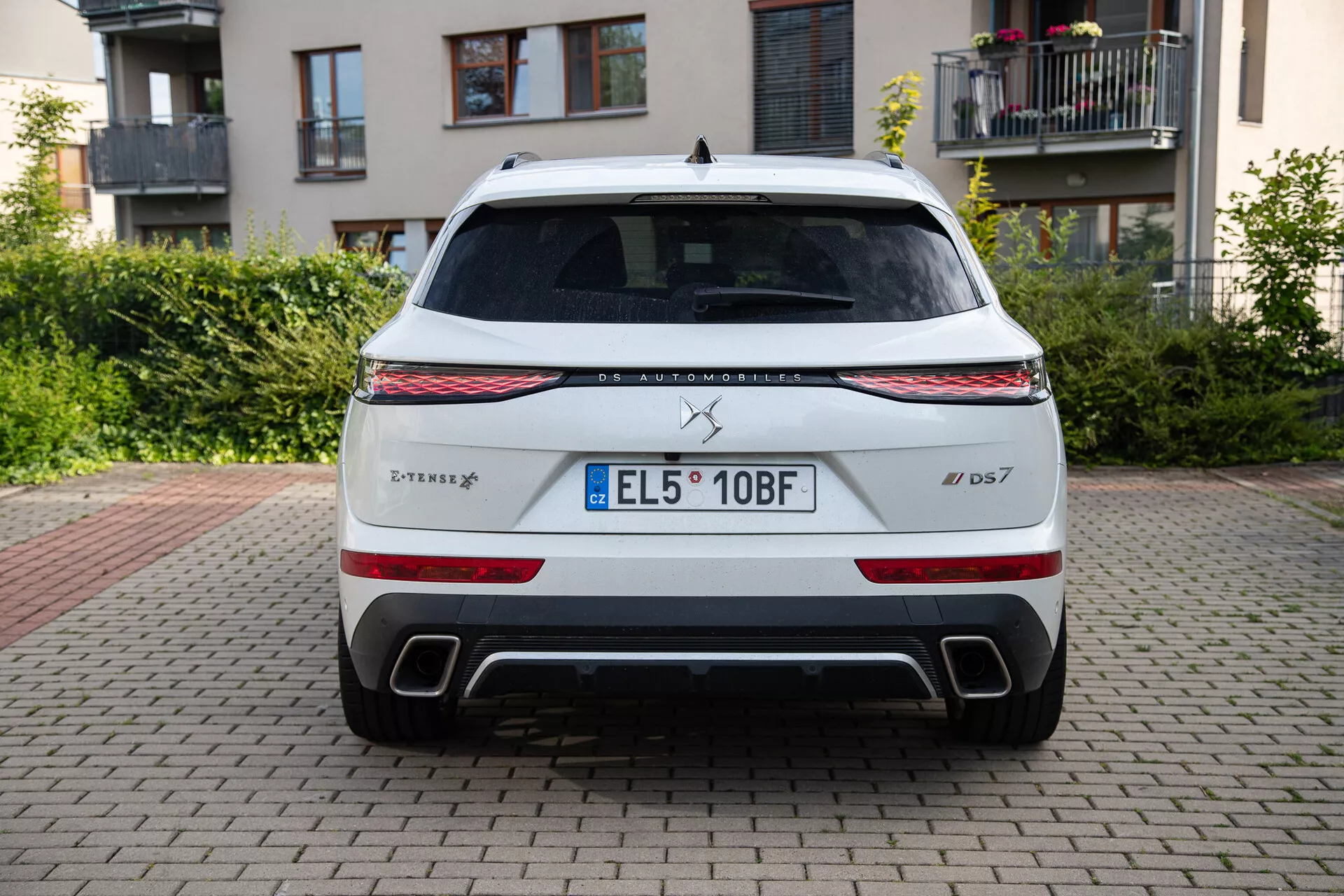 Vue arrière d'une DS Automobiles DS 7 2022 garée sur une surface en briques avec des bâtiments et de la verdure en arrière-plan, présentant une bande lumineuse LED, des feux arrière, des sorties d'échappement doubles et une plaque d'immatriculation de l'UE