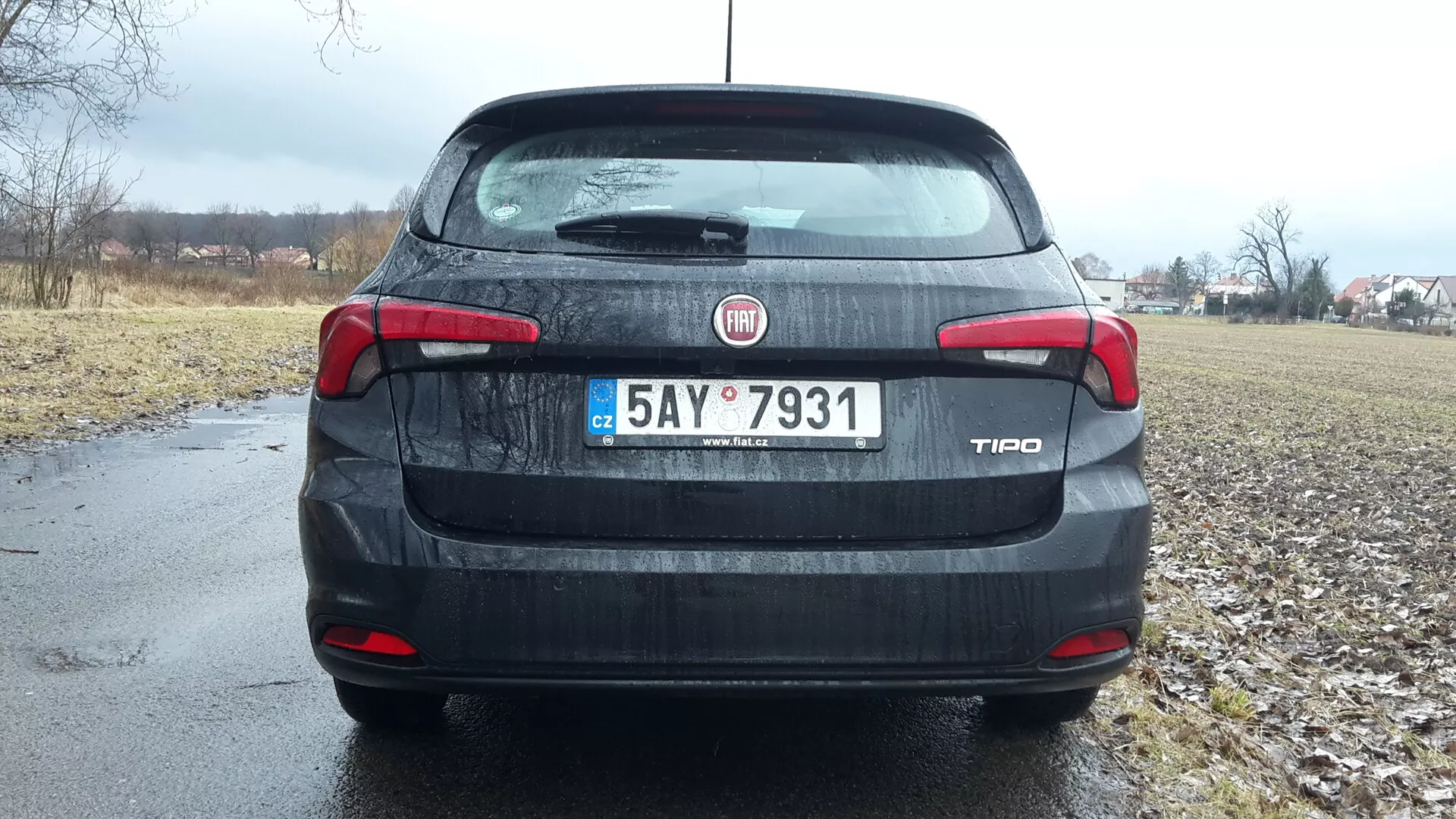 Vue arrière d'une berline Fiat Tipo 2016 sous un ciel couvert, présentant l'identification de la marque et du modèle sur le coffre, des feux arrière et un troisième feu stop détaillés, une plaque d'immatriculation et un aperçu de cadre rural en arrière-plan