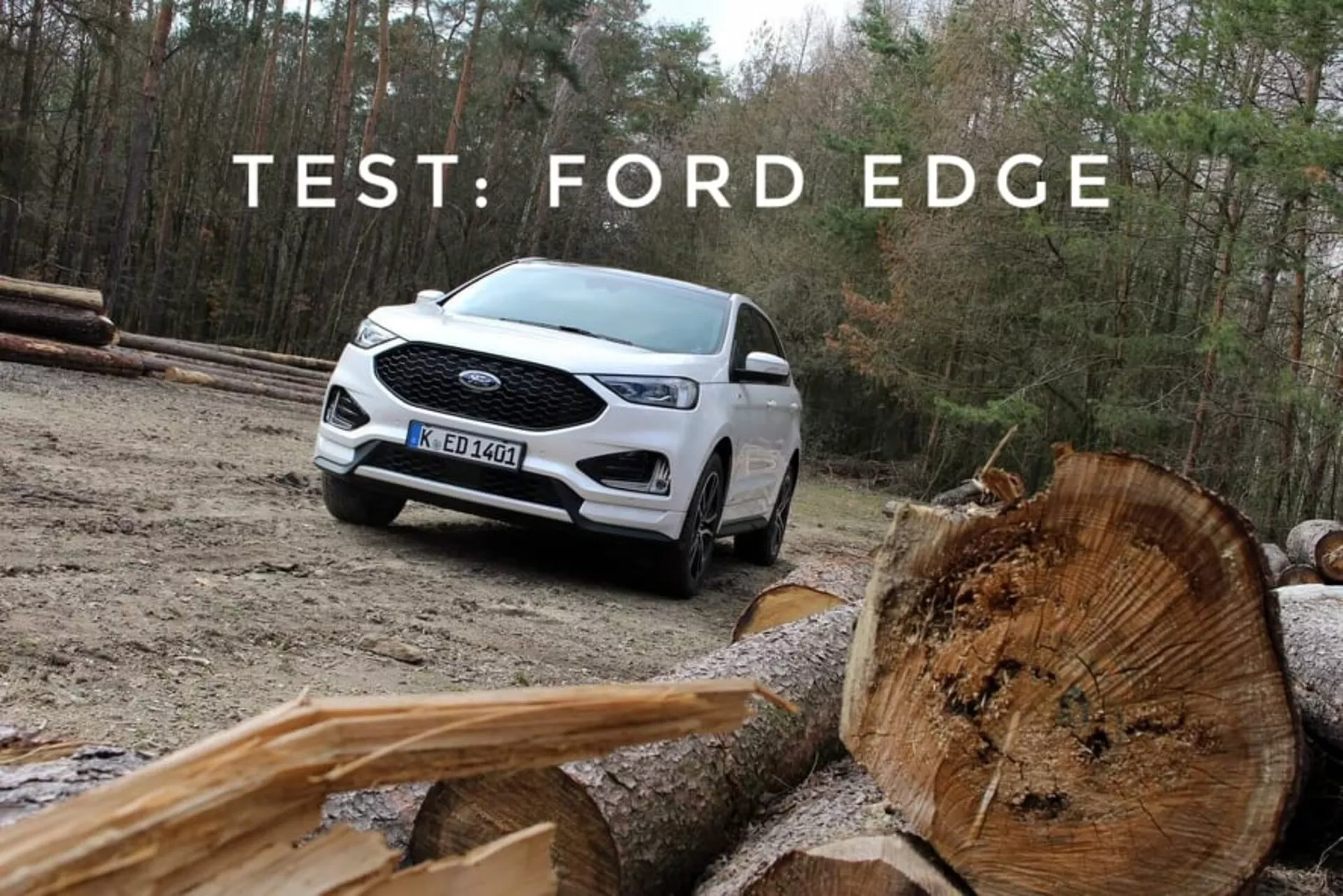 Ford Edge 2018 blanc, démontrant sa capacité tout-terrain avec une vue de trois quarts avant et latérale, mettant en avant les feux de jour à LED, les roues en alliage à multi-rayons, et la peinture bicolore, situé contre un arrière-plan naturel avec des bûches de bois coupées au premier plan