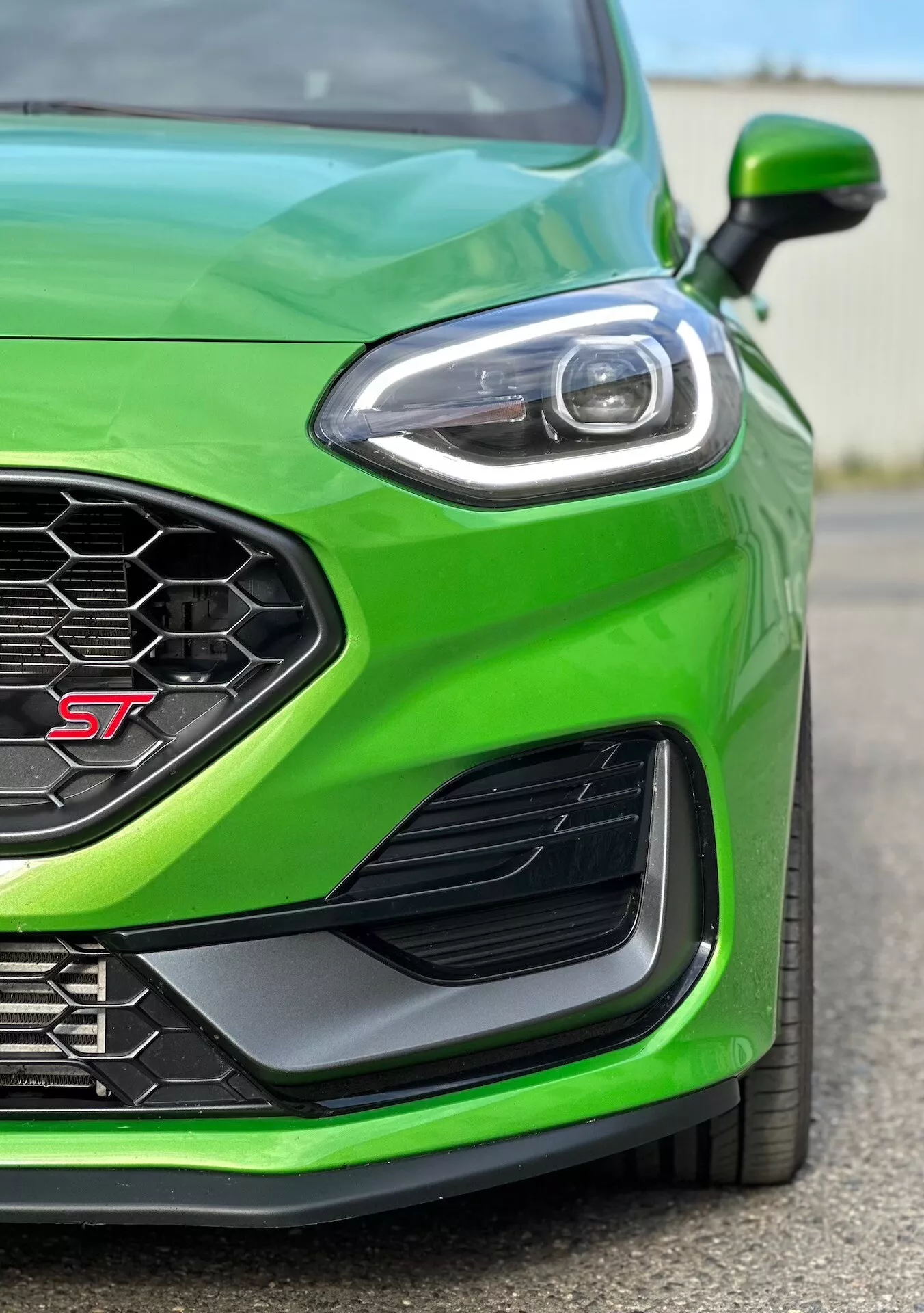 Vue rapprochée du quart avant gauche d'une Ford Fiesta ST 2022 en vert vibrant, mettant en avant la finition sportive, la grille en nid d'abeille pour un flux d'air optimal, le design avancé des phares LED, l'évent latéral aérodynamique et le pare-chocs avant pour une meilleure canalisation de l'air.