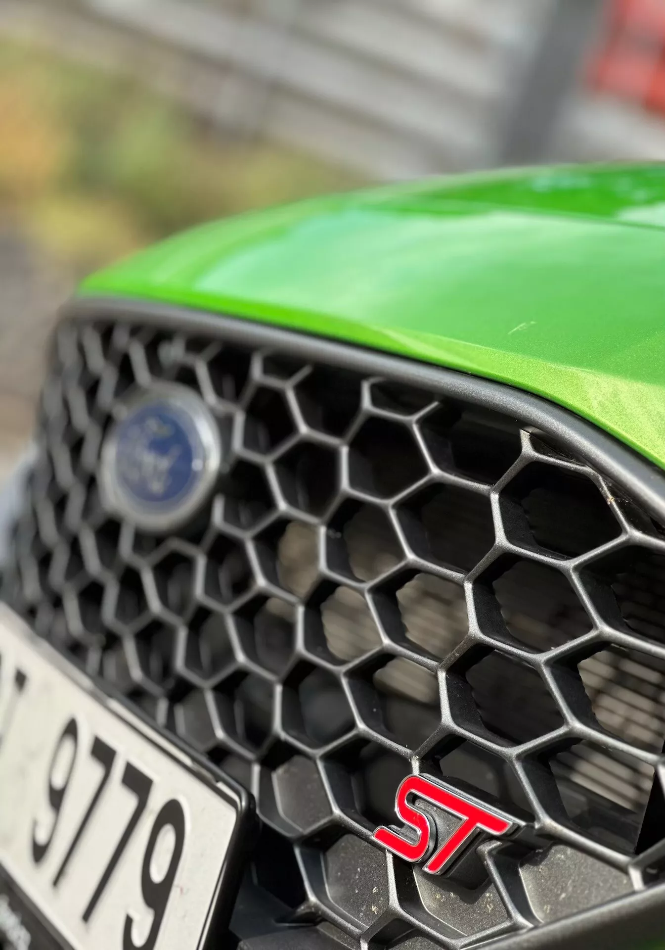 Vue rapprochée d'une partie avant d'une Ford Fiesta ST 2022 montrant la grille hexagonale, l'insigne ST, et le logo de la marque en mise au point nette avec un capot vert, mettant en lumière les caractéristiques de performance et de design