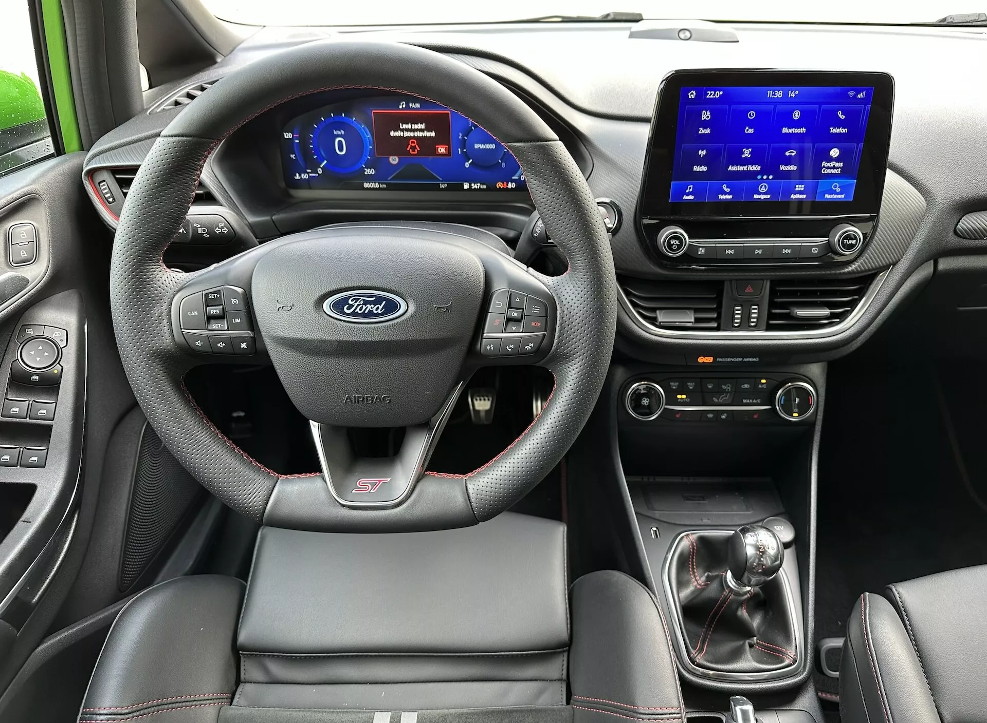 Vue intérieure de la Ford Fiesta 2022 depuis le siège arrière, mettant en avant le volant avec logo Ford et badge ST, tableau de bord numérique, système d'infodivertissement tactile, levier de vitesse manuel et siège conducteur sportif avec coutures contrastées.