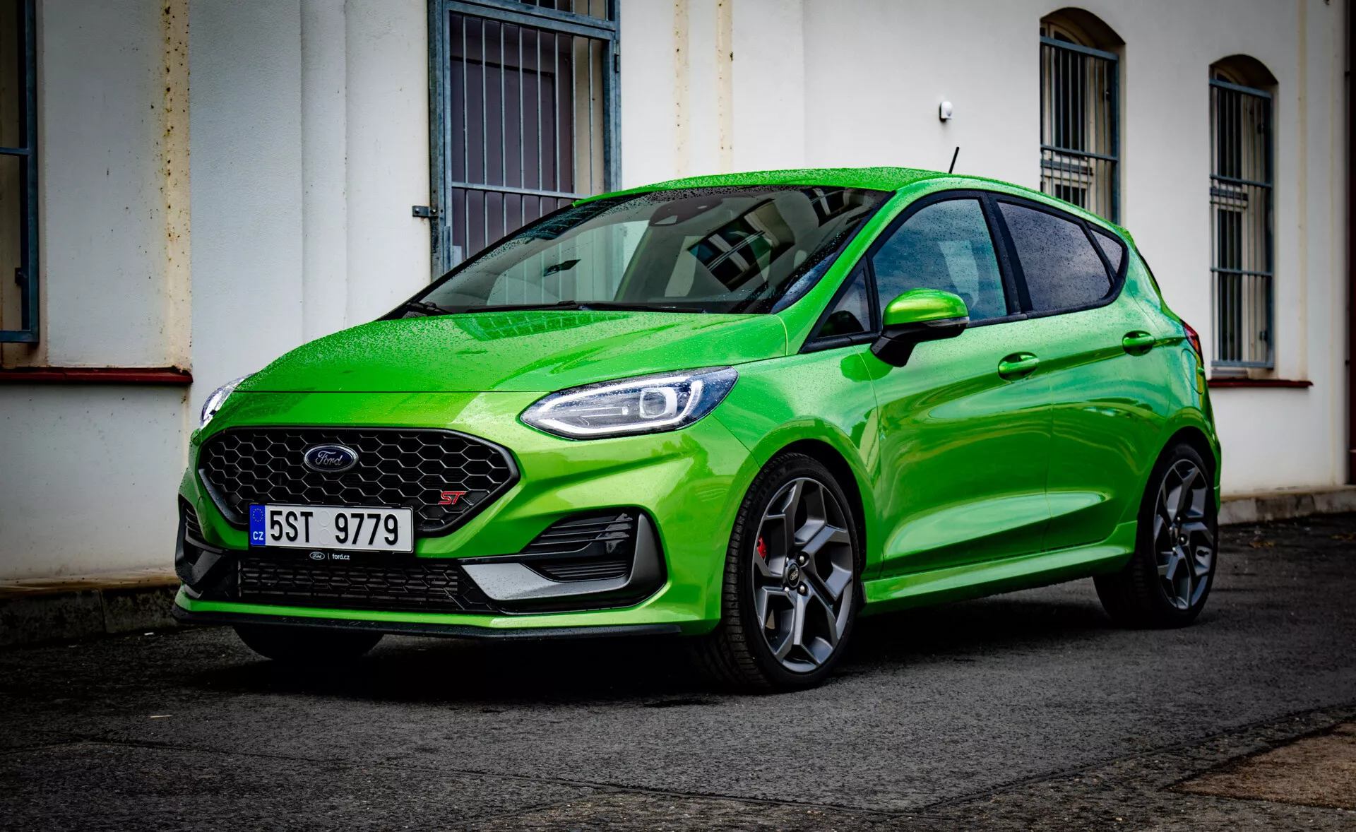 Ford Fiesta 2022 en vert éclatant, présentant son design sportif avec une vue angulaire avant basse de trois quarts mettant en évidence le pare-chocs avant, la grille, les phares, le capot, l'aile, les roues et le reflet du pare-brise dans une rue urbaine après la pluie