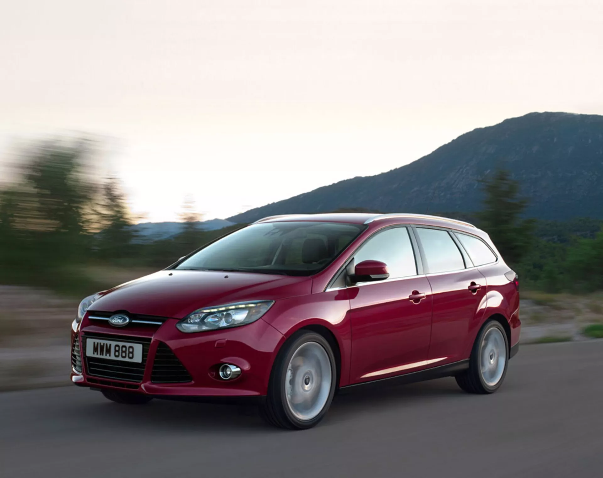 Break Ford Focus rouge 2011 en mouvement sur une route en asphalte, mettant en avant son design moderne, grille avant proéminente avec le logo Ford, phares arrière inclinés, lignes sculptées sur le capot et jantes en alliage, dans des conditions d'éclairage du soir