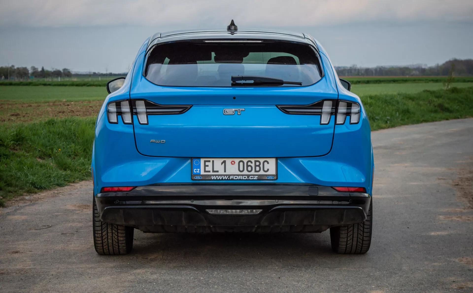 Vue arrière d'une Ford Mustang-Mach-E bleue de 2020 sur une route en asphalte avec des champs d'herbe en arrière-plan, mettant en lumière des caractéristiques clés telles que le logo du fabricant, le nom du modèle, le badge de niveau de finition, la plaque d'immatriculation polonaise, le système d'échappement double, les feux arrière LED modernes, les capteurs de stationnement et le feu stop surélevé.