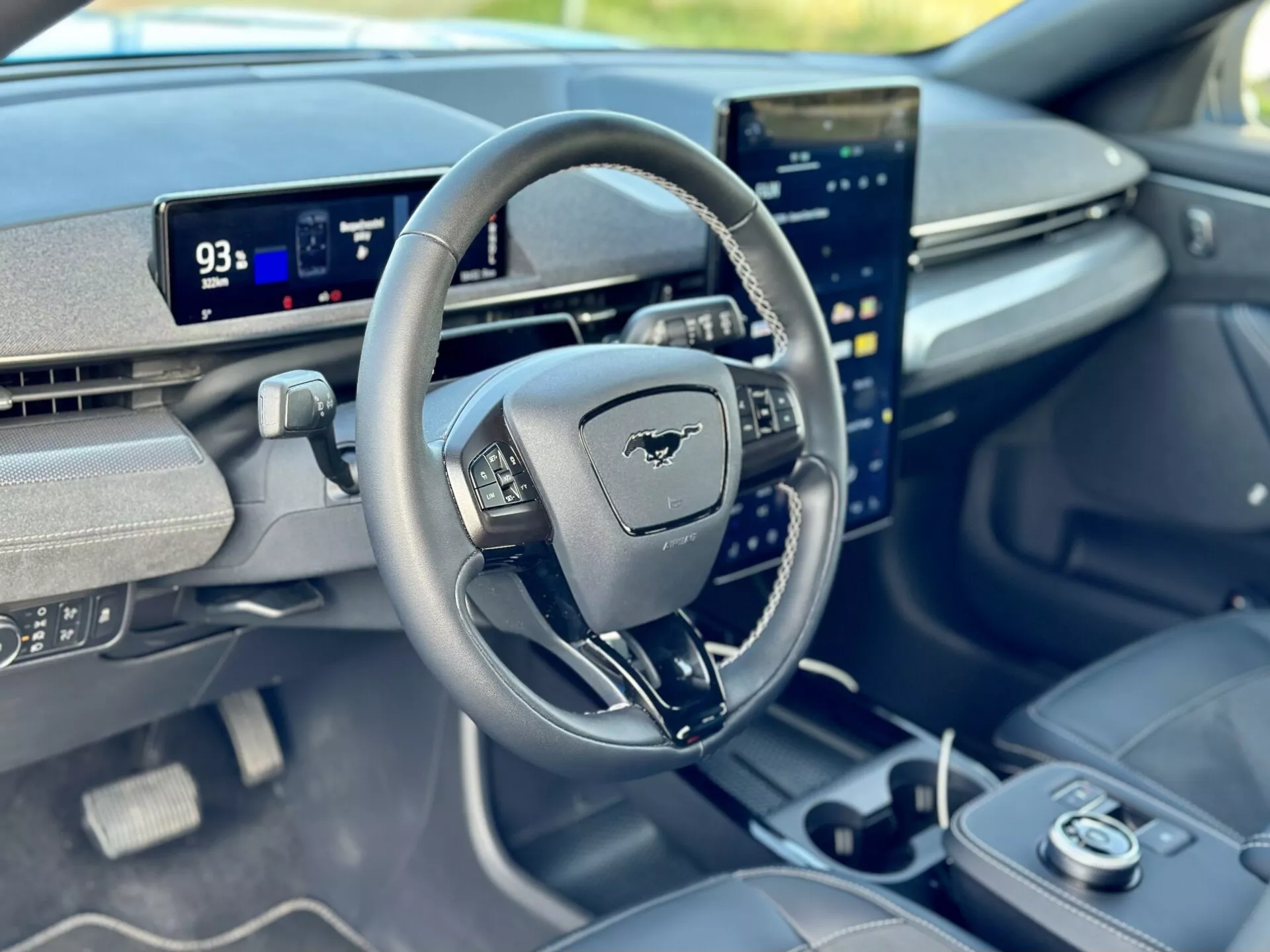 Vue intérieure d'une Ford Mustang Mach-E 2020 se concentrant sur le cockpit du conducteur depuis la perspective du passager avant, présentant un volant avec l'emblème Mustang, un tableau de bord numérique en mode Parking affichant une autonomie de 93 miles, et un écran tactile central orienté verticalement intégré avec des capacités d'infodivertissement avancées.