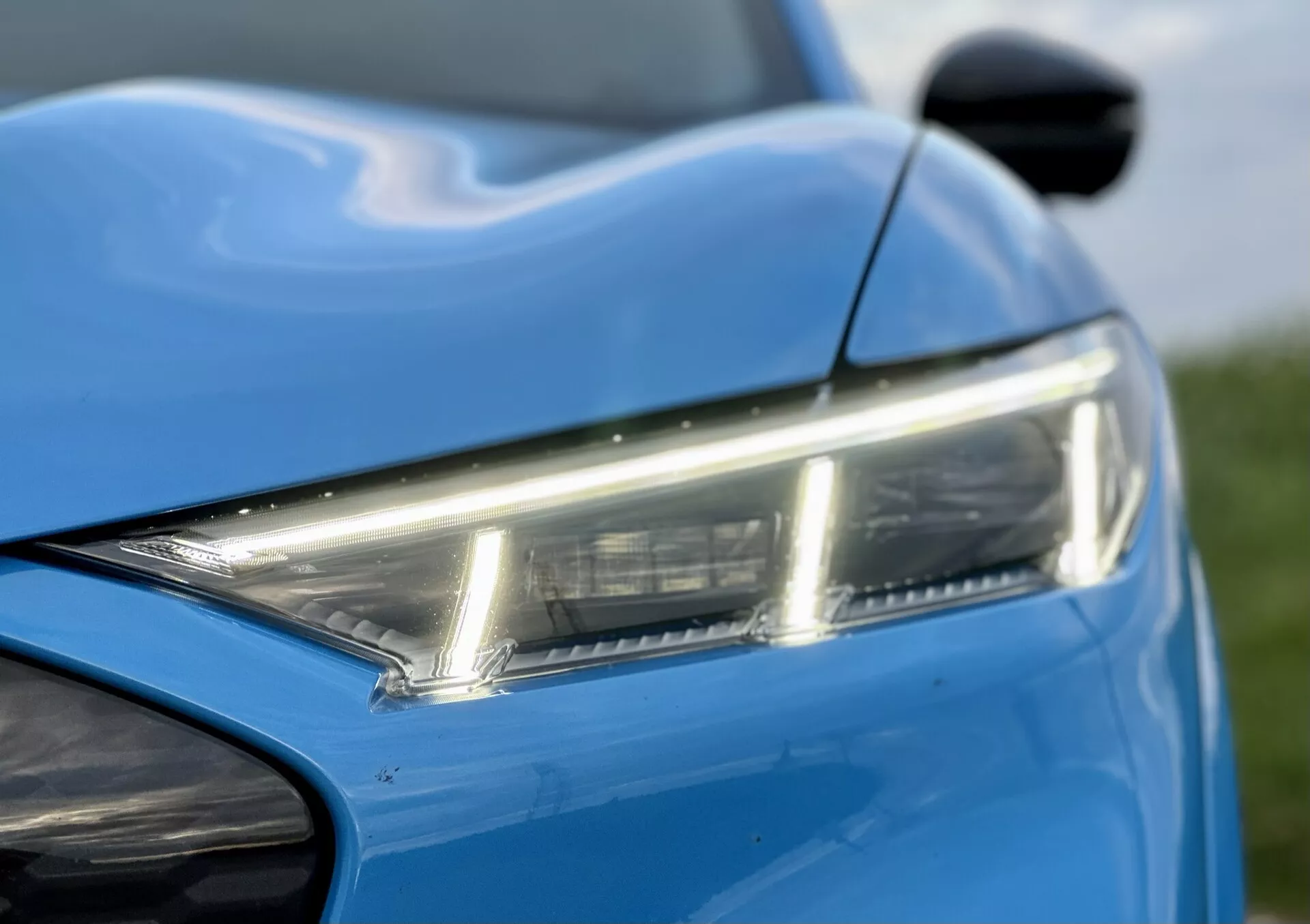 Vue rapprochée de la section avant gauche du Ford Mustang-Mach-E 2020, mettant en évidence le phare LED élégant, la peinture bleue vibrante et les lignes dynamiques de la carrosserie, avec un aperçu du rétroviseur latéral et du pare-choc avant, démontrant le design moderne et l'esthétique.